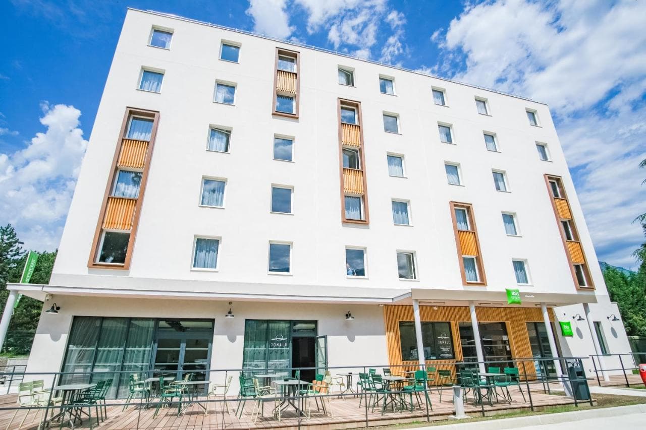 Ibis Albertville hotel exterior with white facade, wooden accents, and outdoor seating under blue sky.