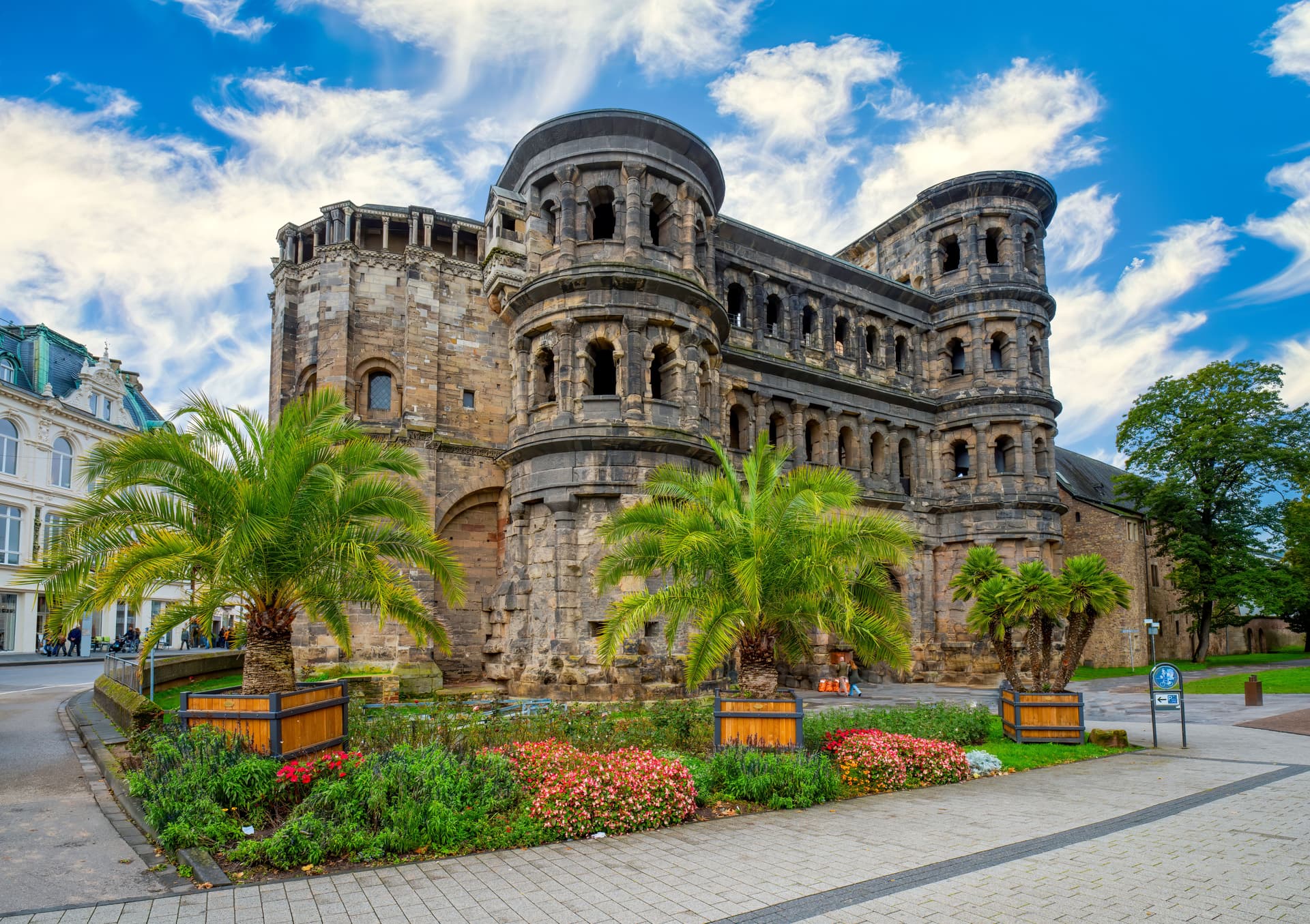 Porta Nigra Roman gate with palm trees and flower beds under a blue, cloudy sky.