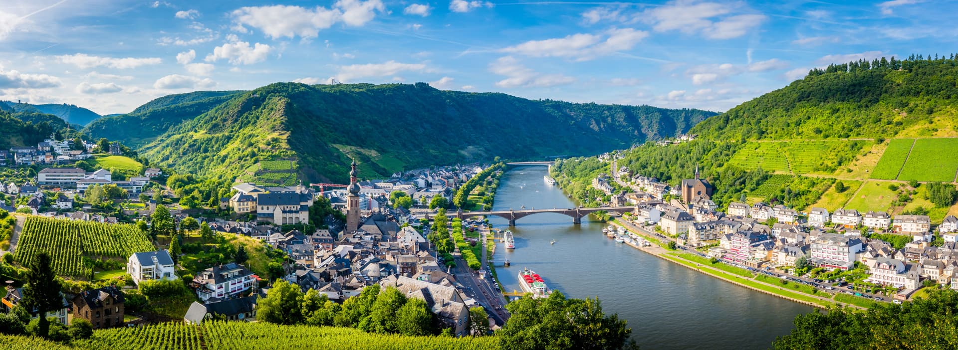 Panoramic view of Trier area town nestled between steep, vine-covered hills and a wide river under a blue sky.