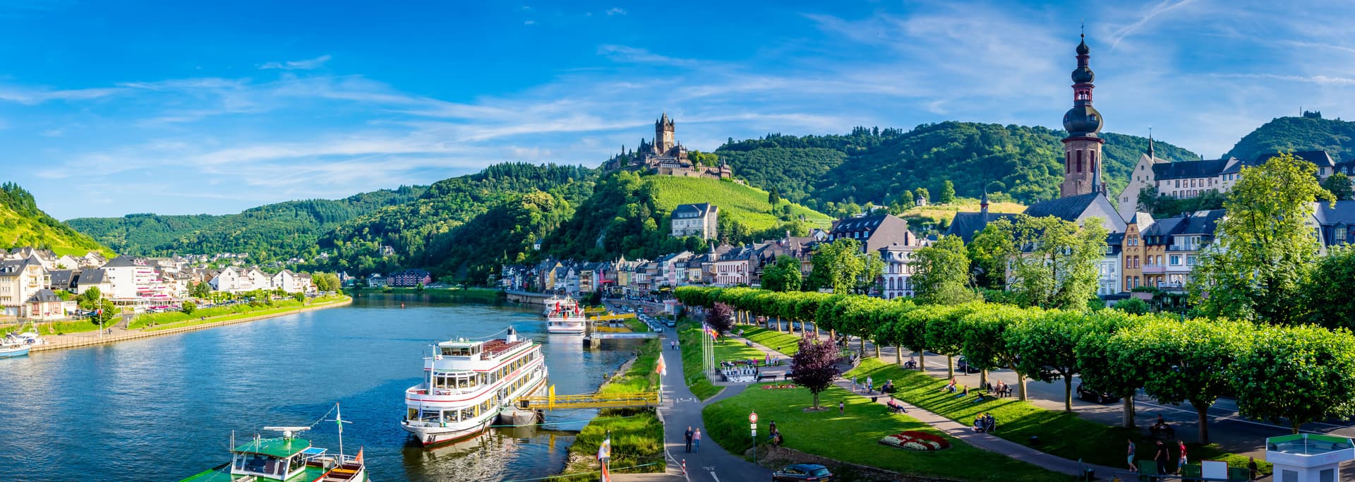 Cochem waterfront with Moselle River, Reichsburg Castle on hill, and church spire under blue sky.