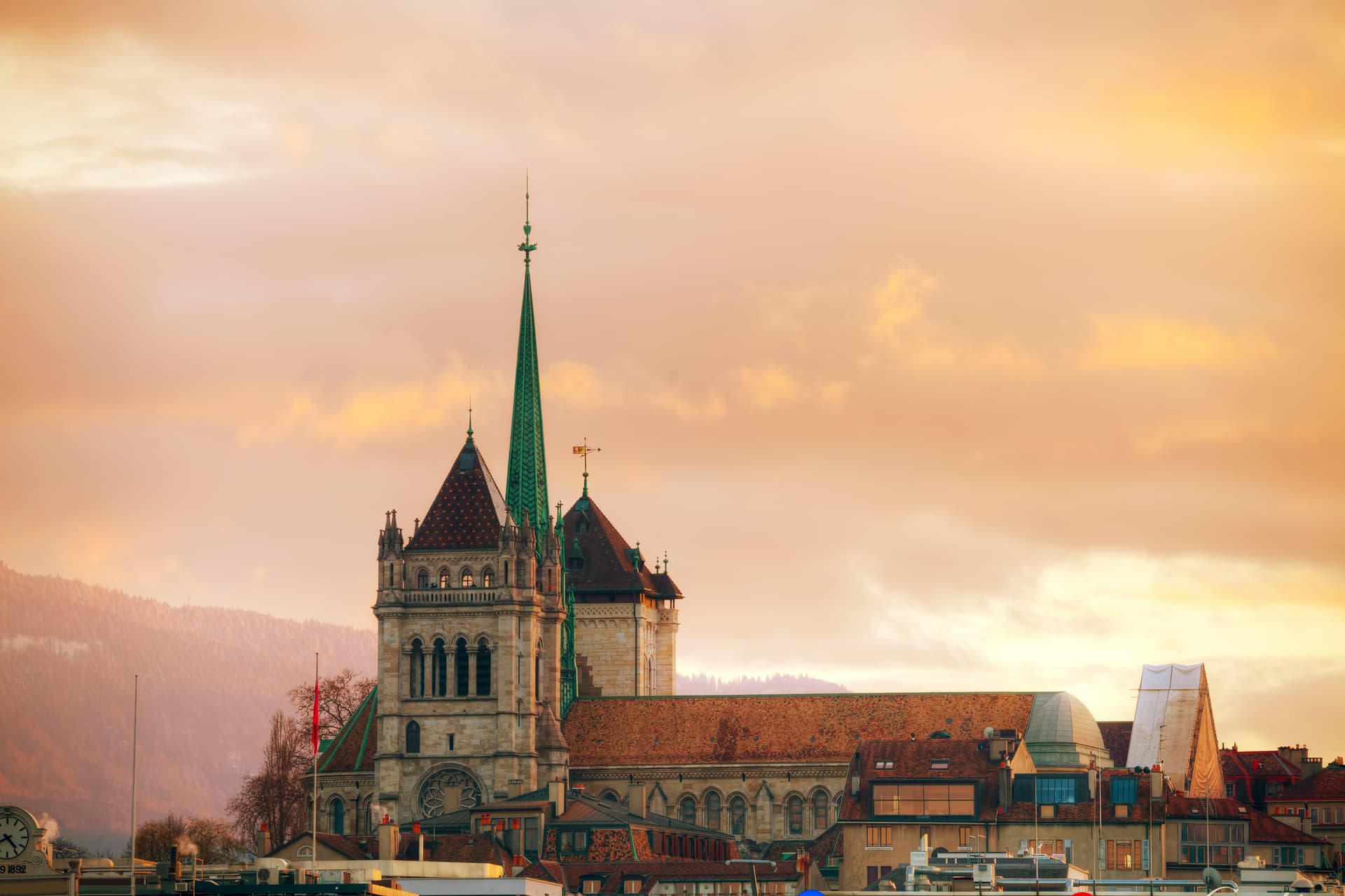 St-Pierre Cathedral spire rising above Geneva cityscape at sunset with mountains in background.