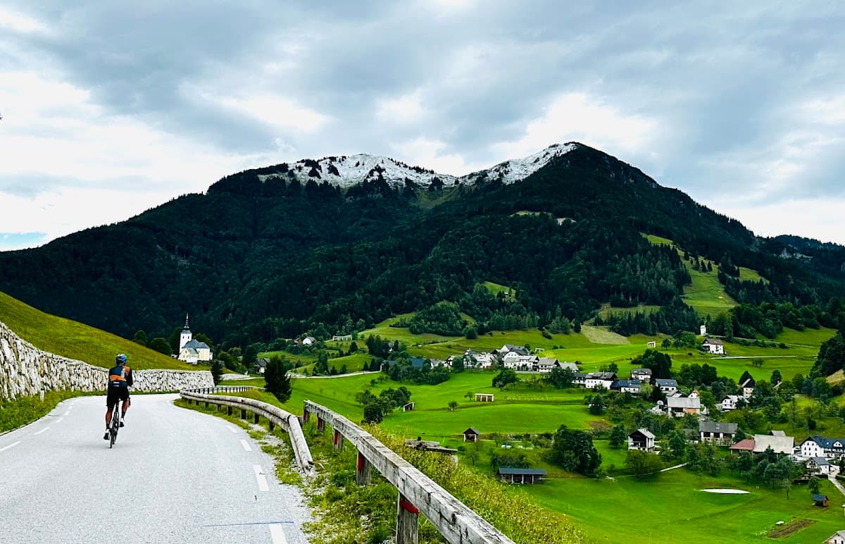 Cyclist riding on mountain road overlooking green valley and village with snow-capped peaks