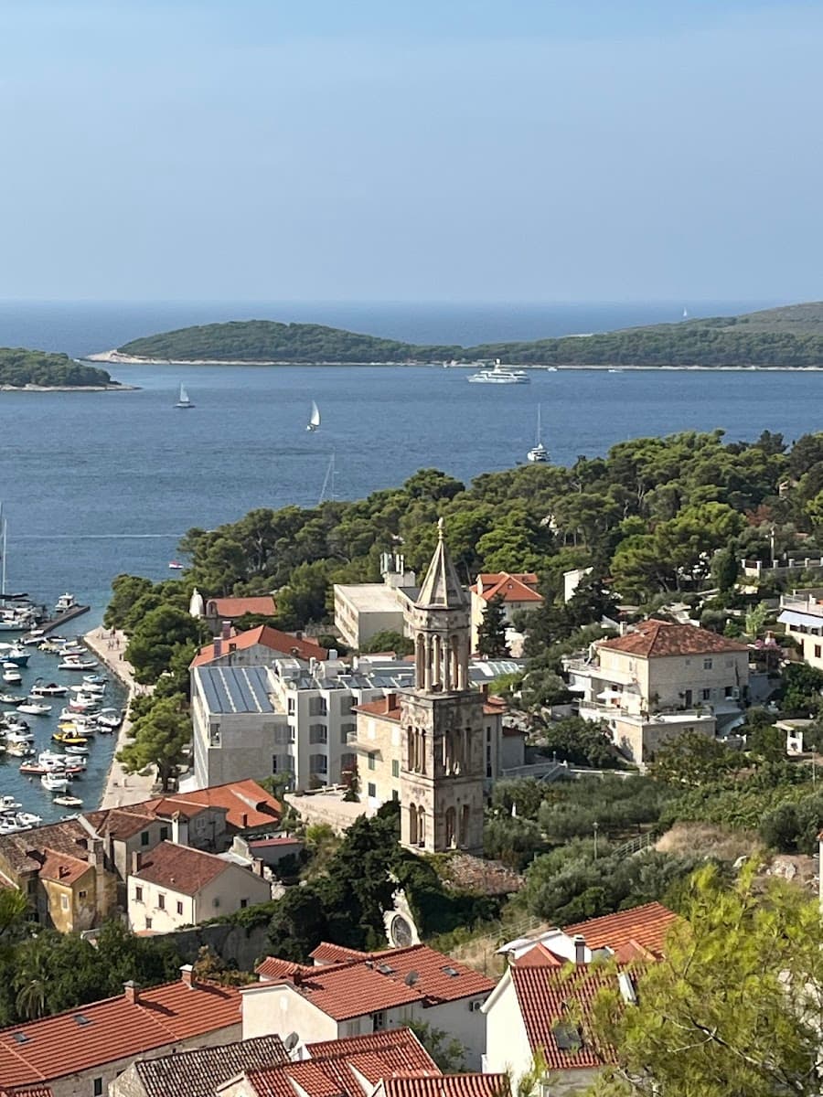 Coastal town with terracotta roofs, a stone bell tower, and boats in the harbor under a clear sky.