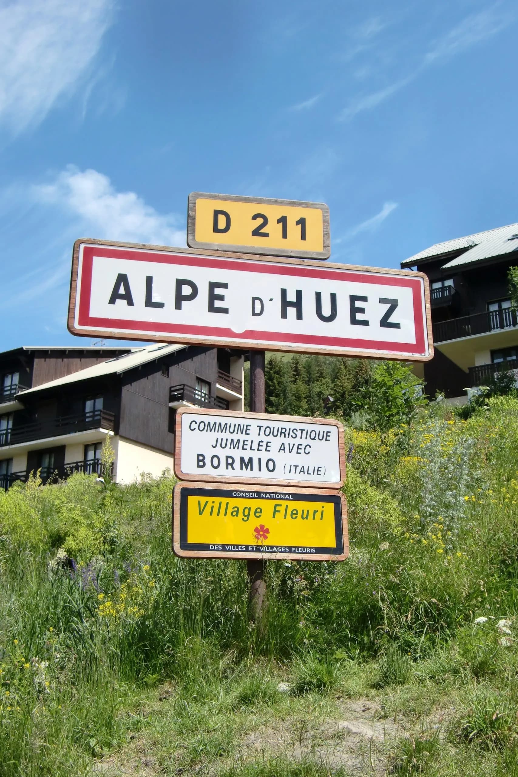 Alpe d'Huez road sign with D 211 marker, Bormio twinning, and Village Fleuri award.
