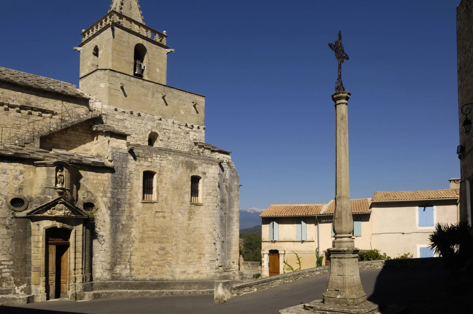 Stone church, calvary cross, and houses under clear sky in Venasque, Provence, France.