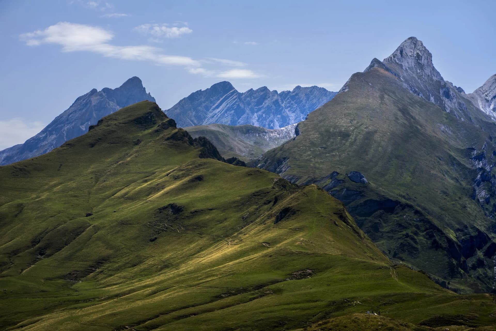 Green rolling mountain slopes beneath rugged, rocky peaks under a blue sky in the Pyrenees.