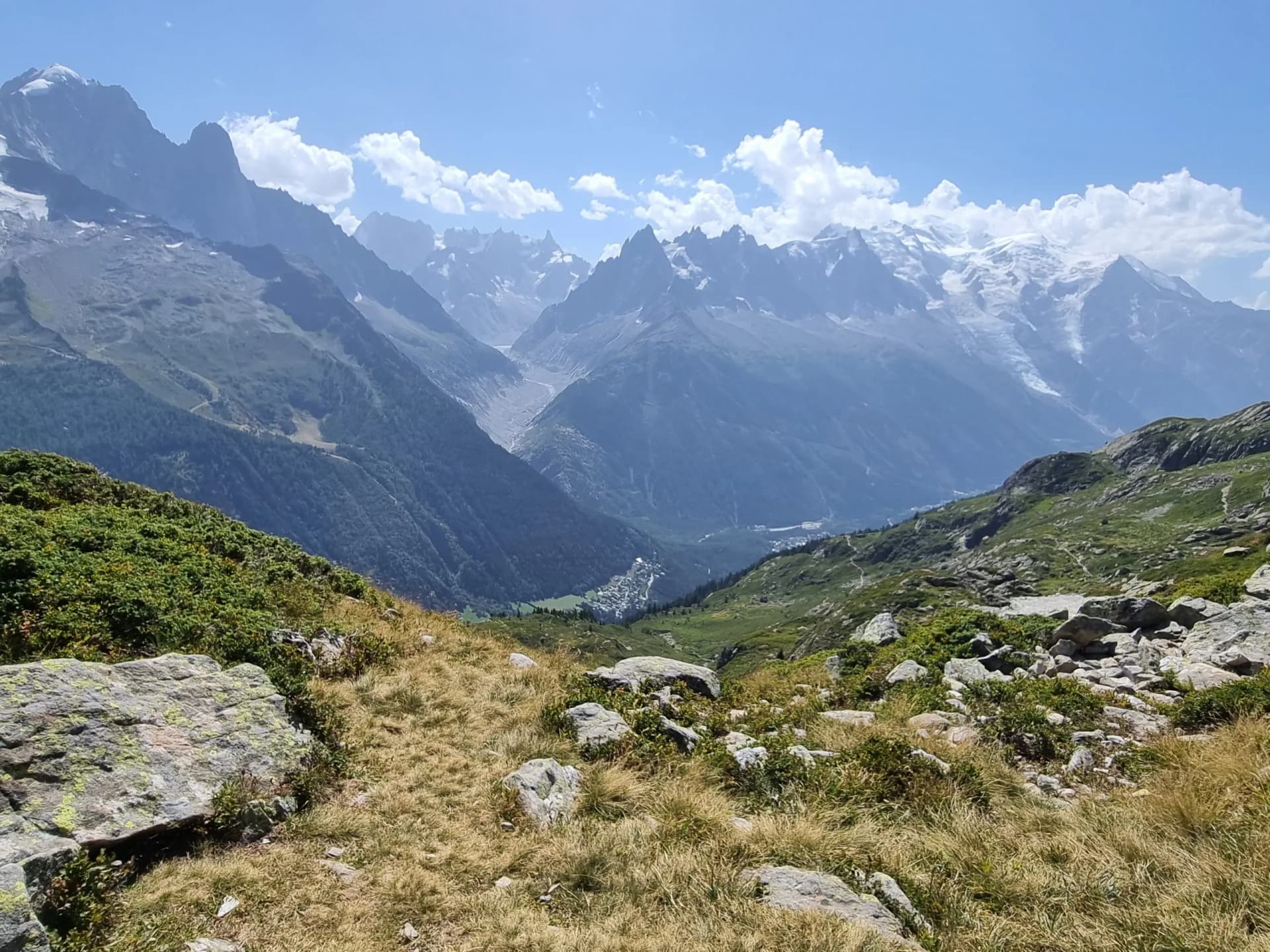 Alpine landscape with rocky foreground, valley, and snow-capped mountains under blue sky.