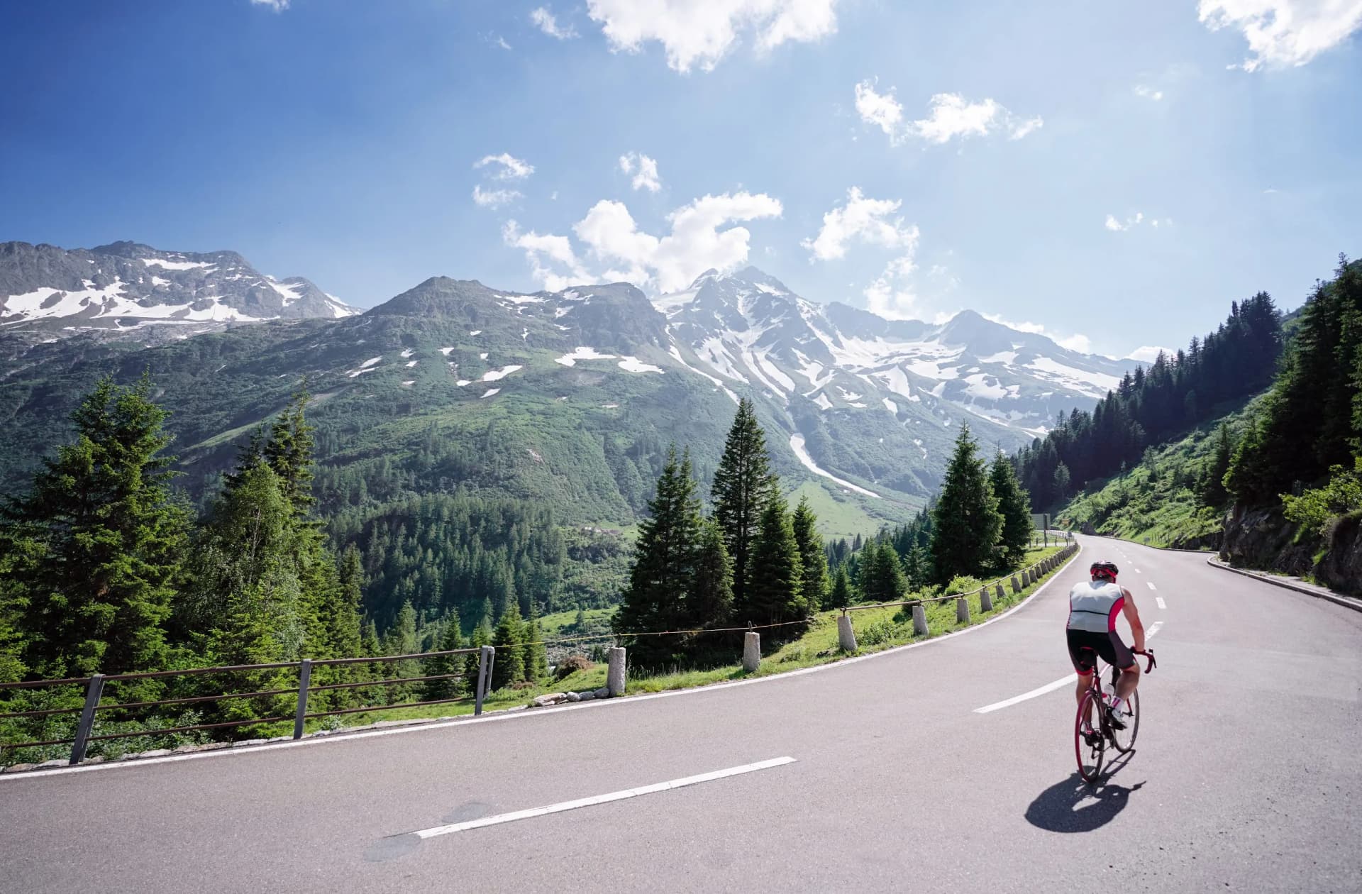 Cyclist riding on mountain road with snow-capped peaks and green forests.