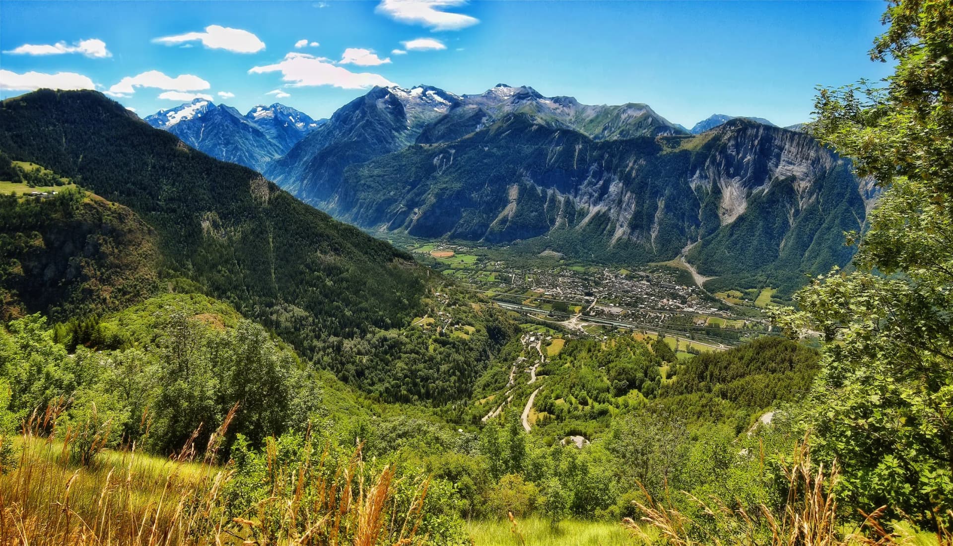 Alpine valley town nestled among green mountains with snow-capped peaks under a blue sky.
