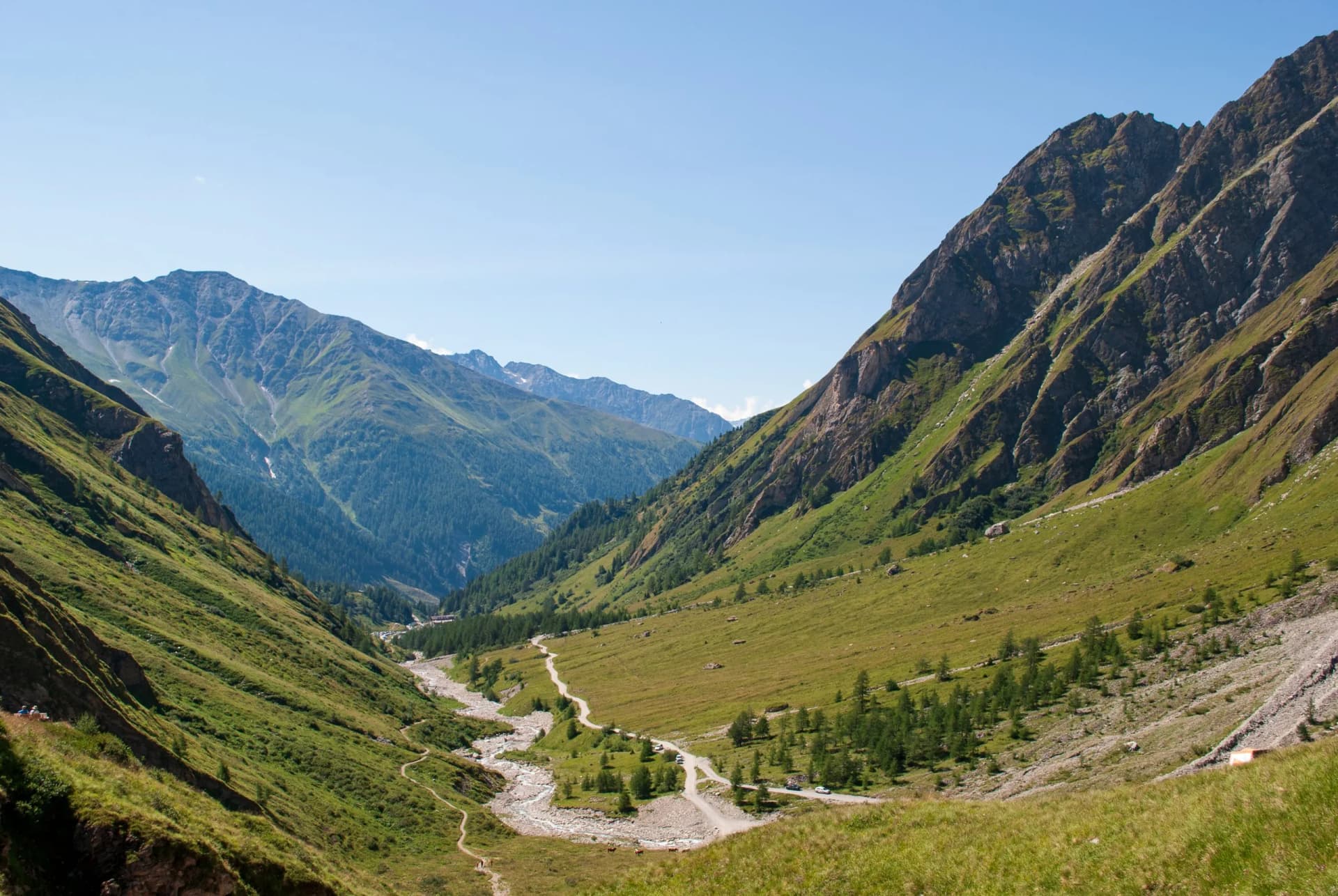 Alpine valley with steep green slopes, a rocky riverbed, and a winding dirt road in summer.