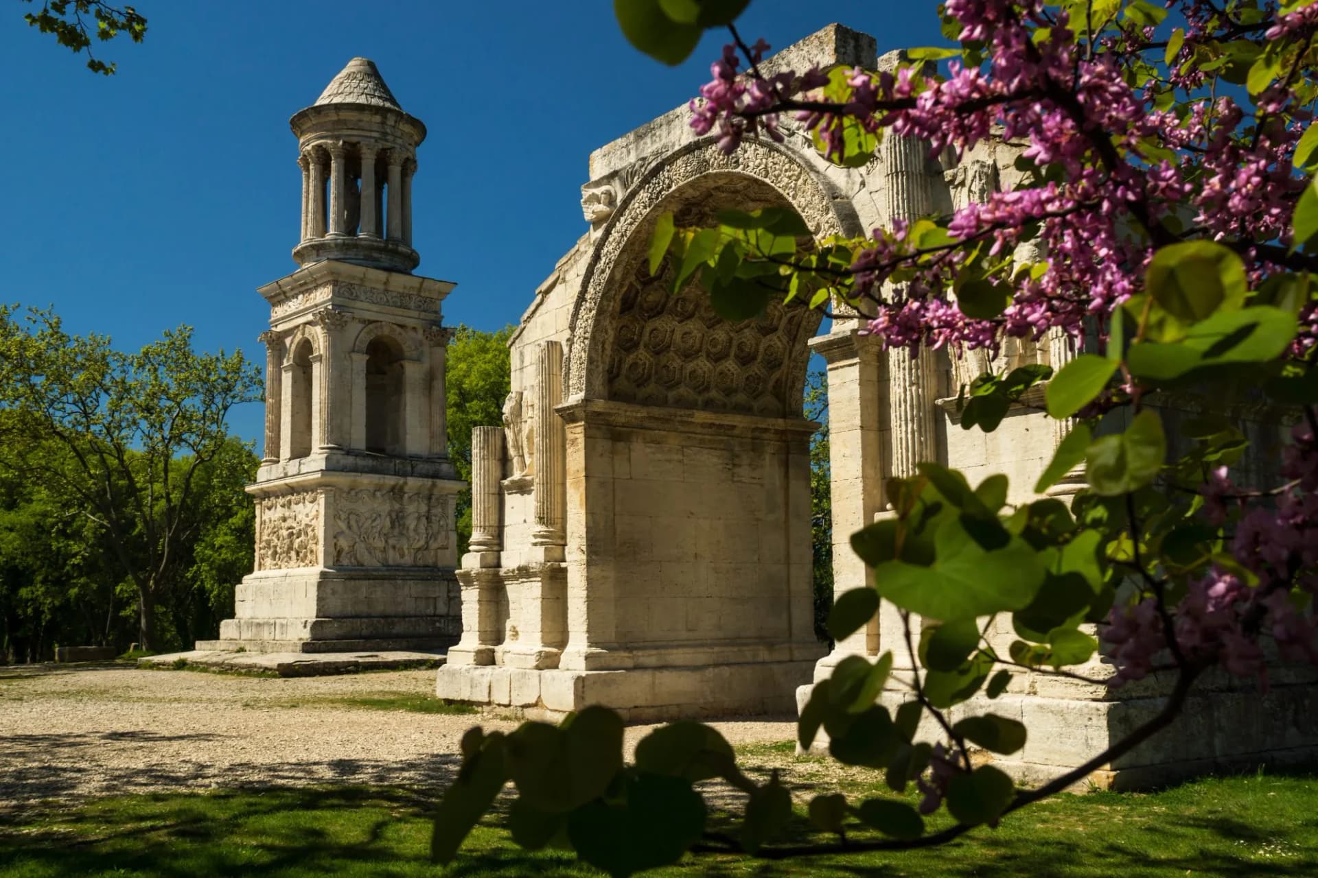 Historic site of Glanum ruins with a funerary monument and arch, framed by spring blossoms.