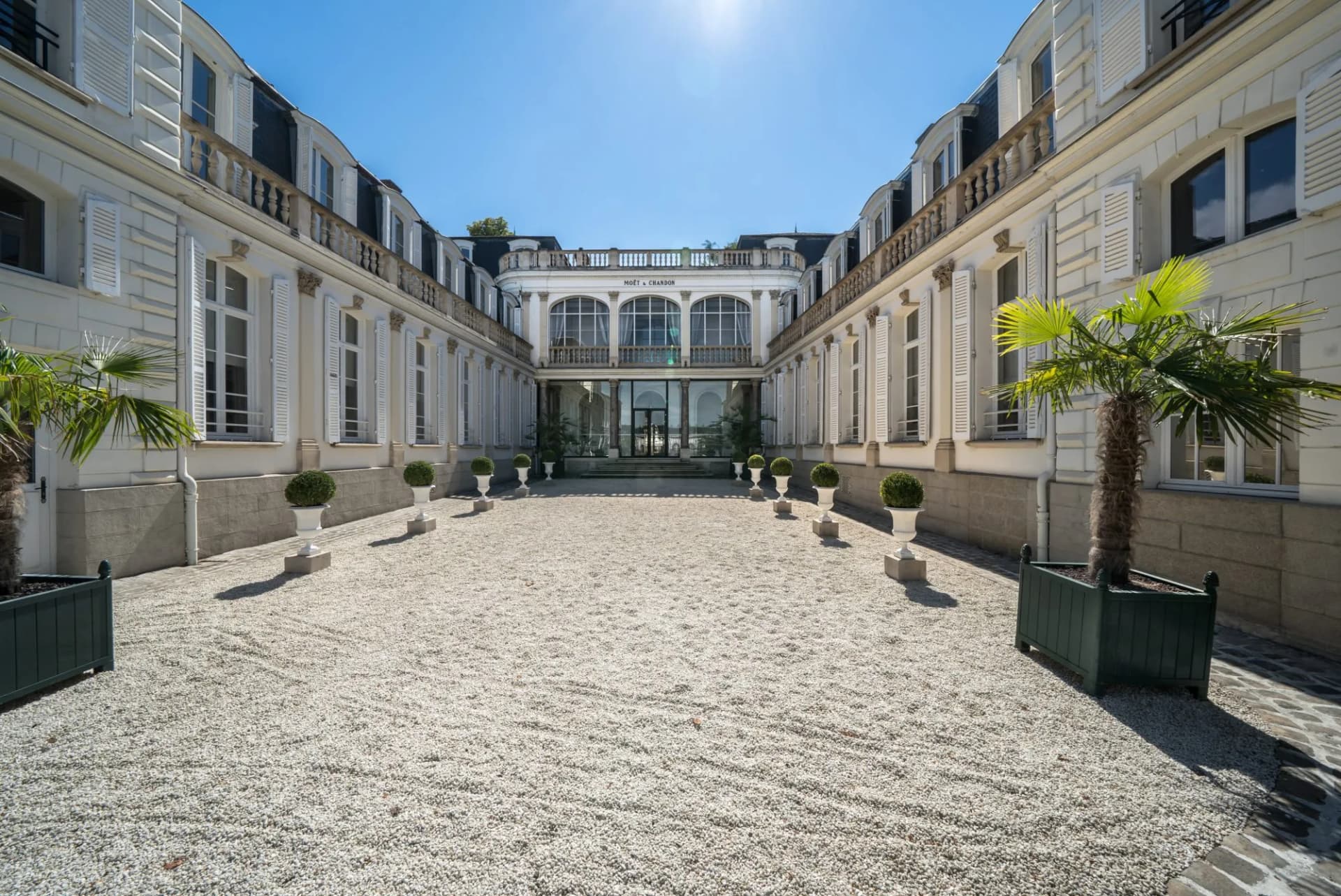 Courtyard of Moët & Chandon house in Epernay with white gravel and potted palms under bright sun.