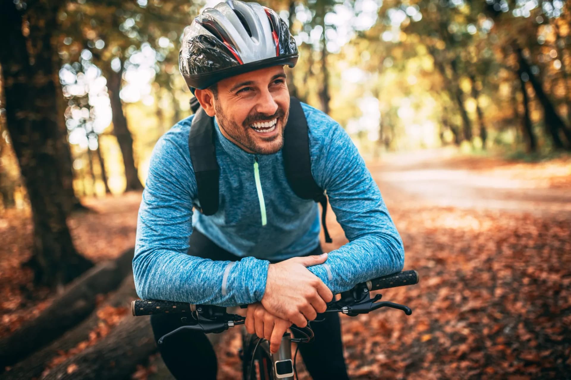 Cyclist wearing a helmet smiles while resting on handlebars in an autumn forest path.