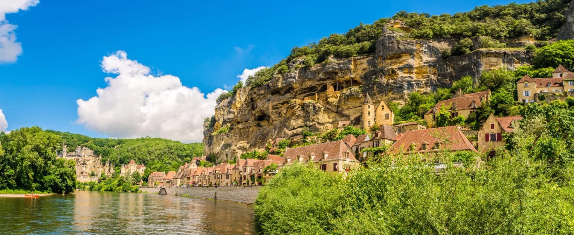Village houses nestled against a cliffside above the Dordogne River under a blue sky.