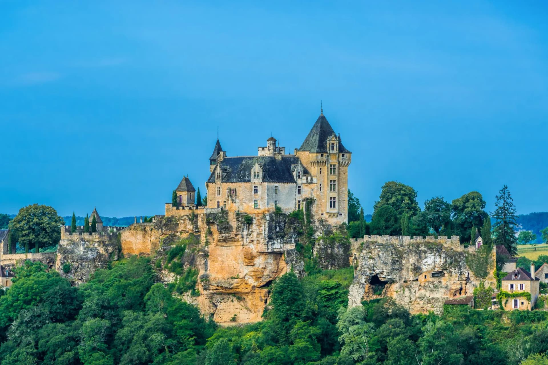 Souillac Castle perched atop a cliff with lush green trees below under a bright blue sky.