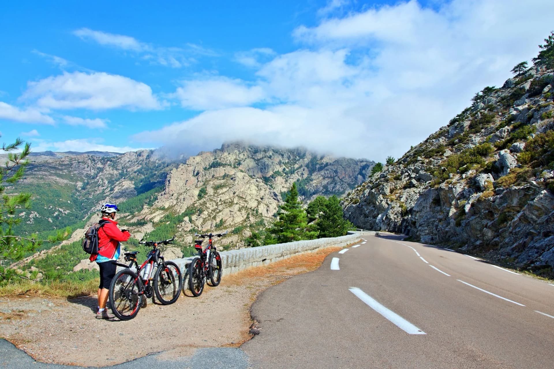 Cyclist with mountain bikes resting by winding road overlooking Corsica mountains near Col de Bavella.