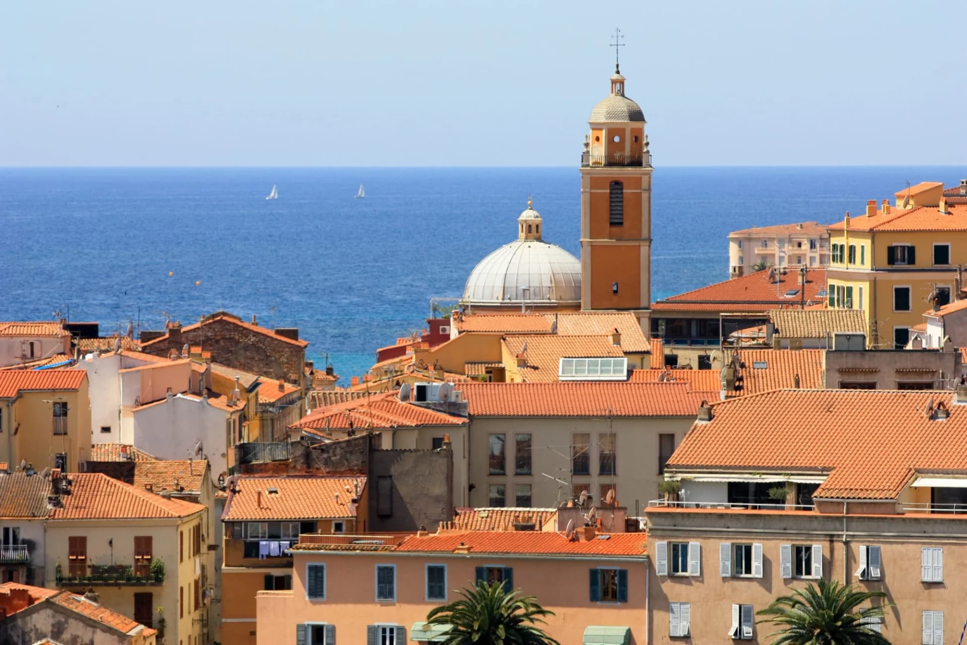 Orange-roofed old town buildings with a church tower overlooking the blue sea and distant sailboats in Ajaccio, Corsica.