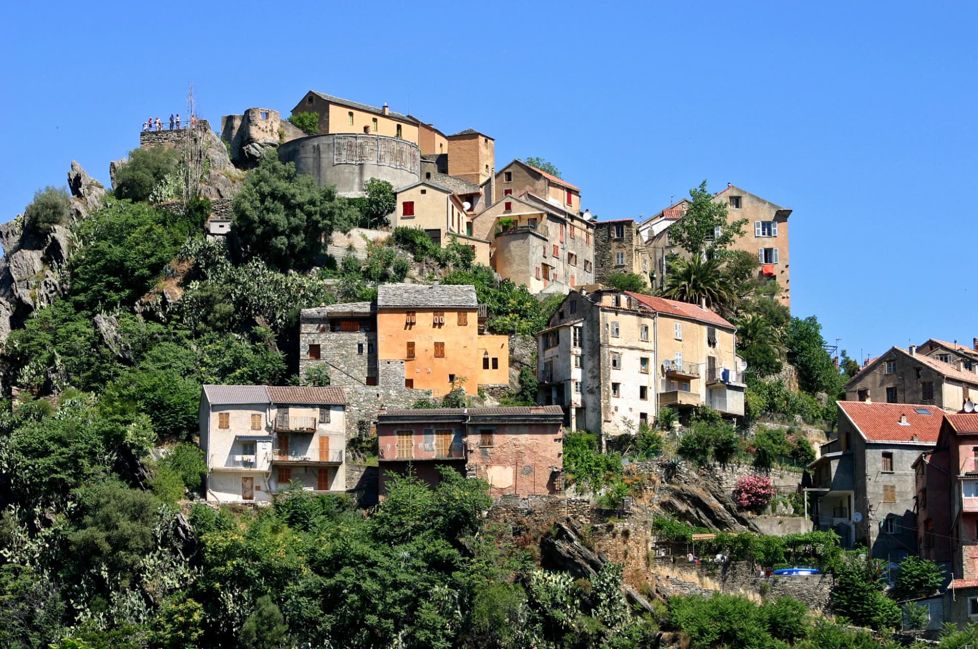 Hillside village with stone houses nestled in lush greenery under a clear blue sky, Citadel of Corte.