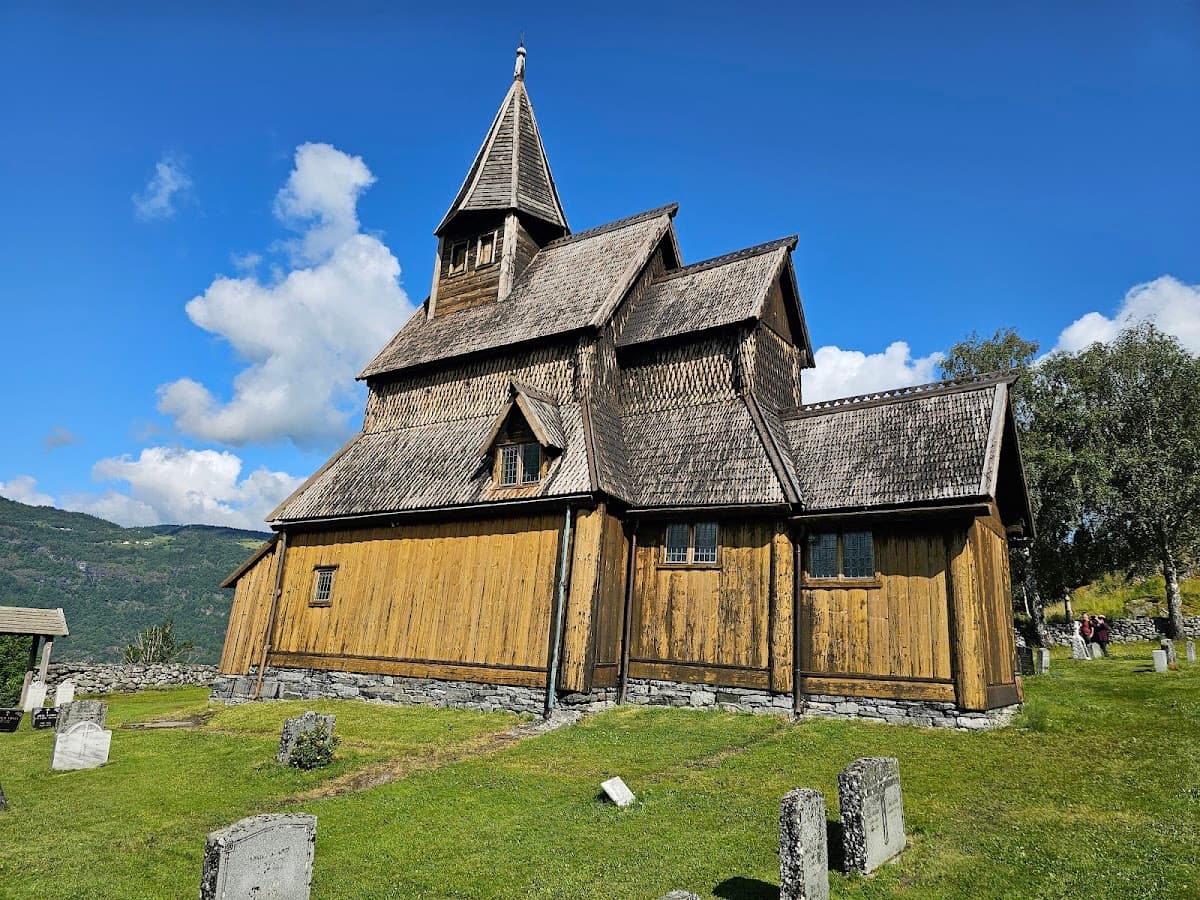Wooden stave church with shingled roof on green grass next to a graveyard, mountains in background.