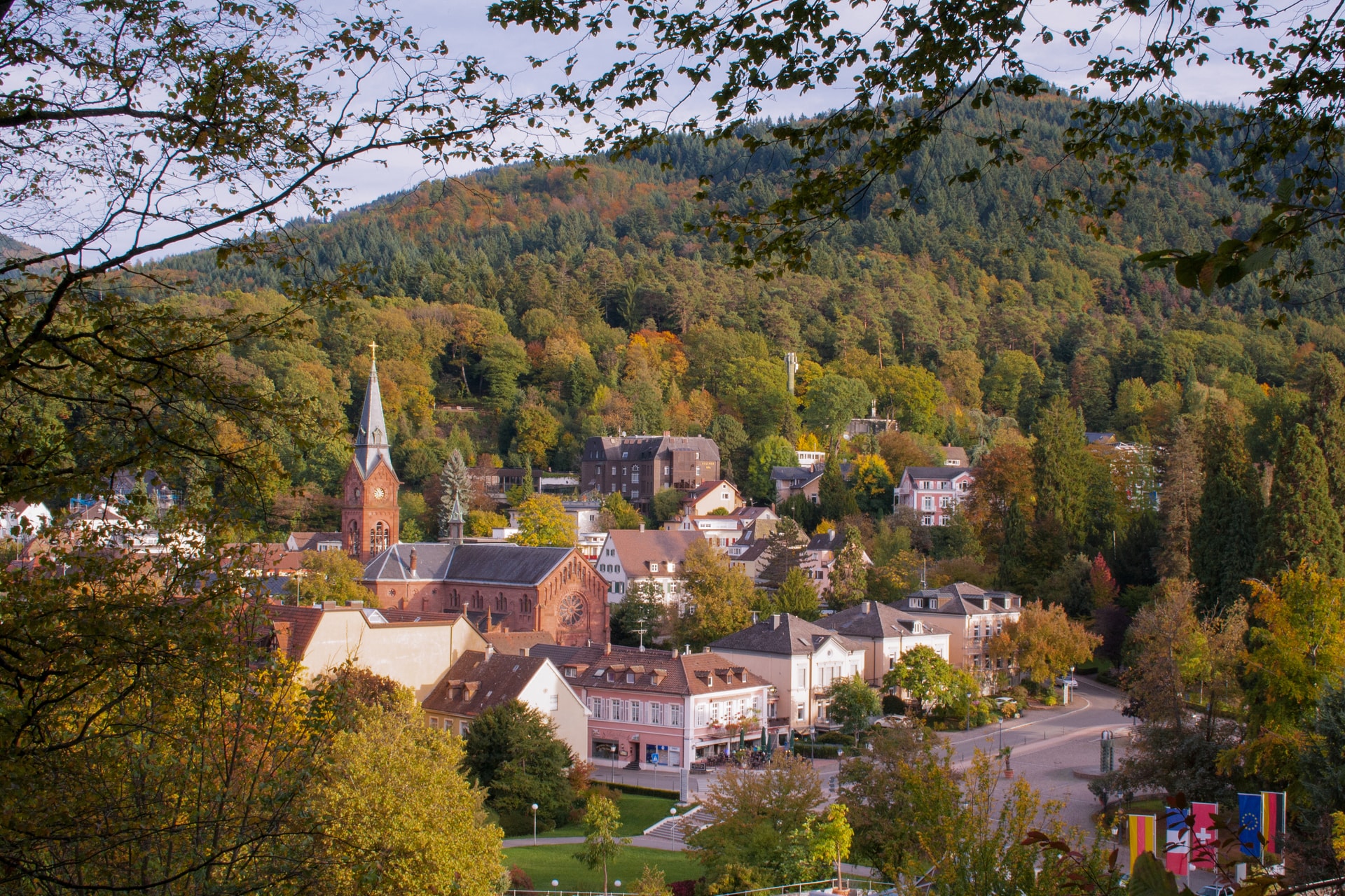 Town with red church spire nestled in autumn forest hills, viewed through tree branches.