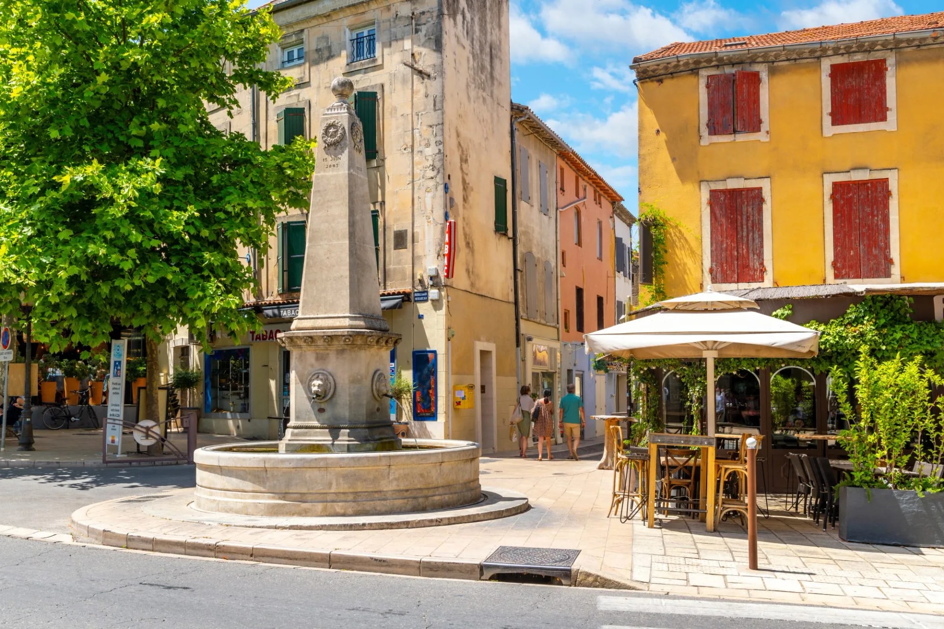 Stone fountain in town square with colorful buildings and outdoor cafe in Saint-Remy-de-Provence.