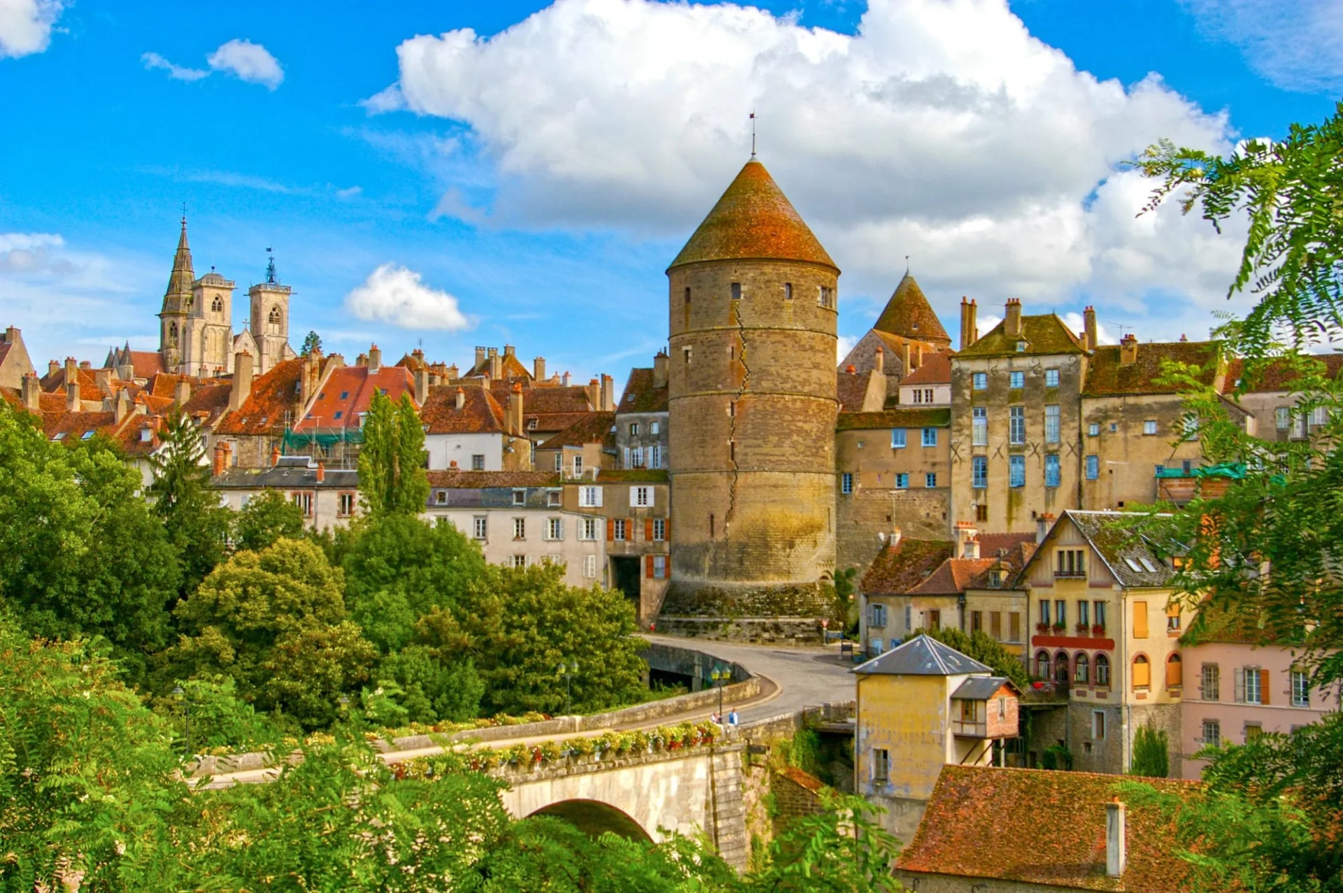 Historic town of Beaune with medieval tower, terracotta roofs, and lush green trees under blue sky.