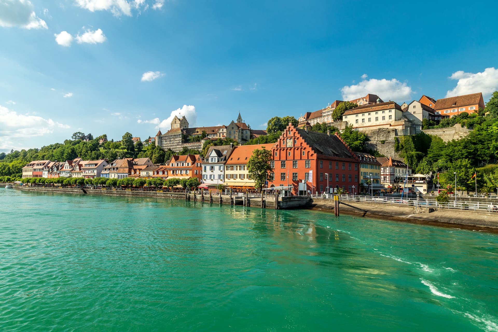 Waterfront of Constance with colorful historic buildings and a castle on a green hill under blue sky.