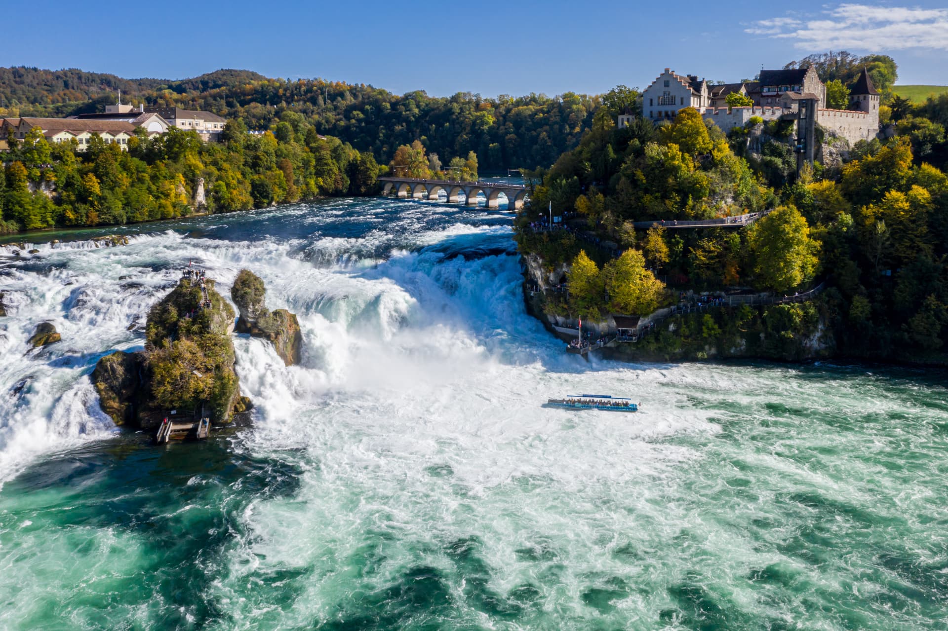 Powerful Rhein Falls with churning turquoise water, a boat tour below, and a castle on the forested cliff.