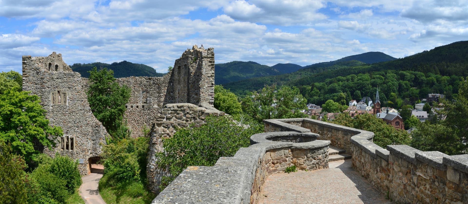 Stone ruins of Burg Badenweiler overlooking a town and forested hills under a cloudy sky.