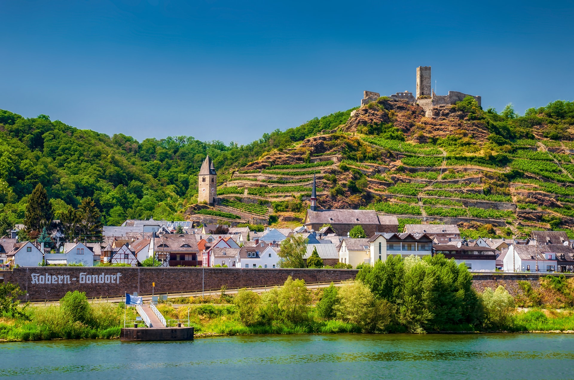 Kobern-Gondorf village on riverbank below terraced vineyards and hilltop castle ruins.