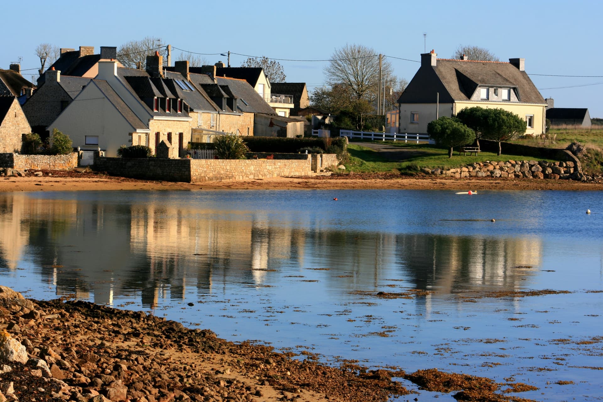 Houses reflected in calm water near a rocky shore in the hamlet of Moustoir, Brittany.