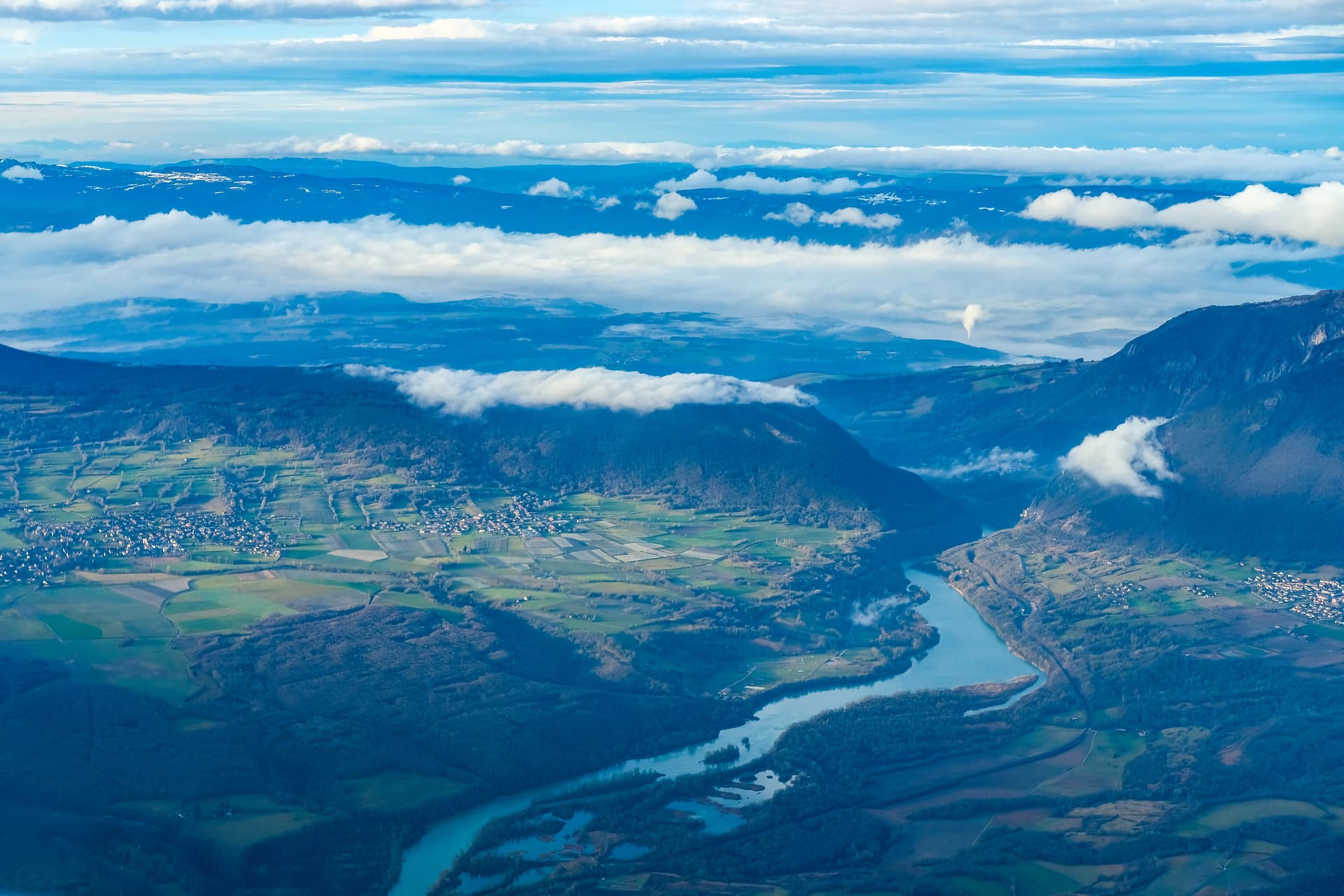 Aerial view of the Rhone River winding through valleys with green fields and villages below low clouds near Geneva.