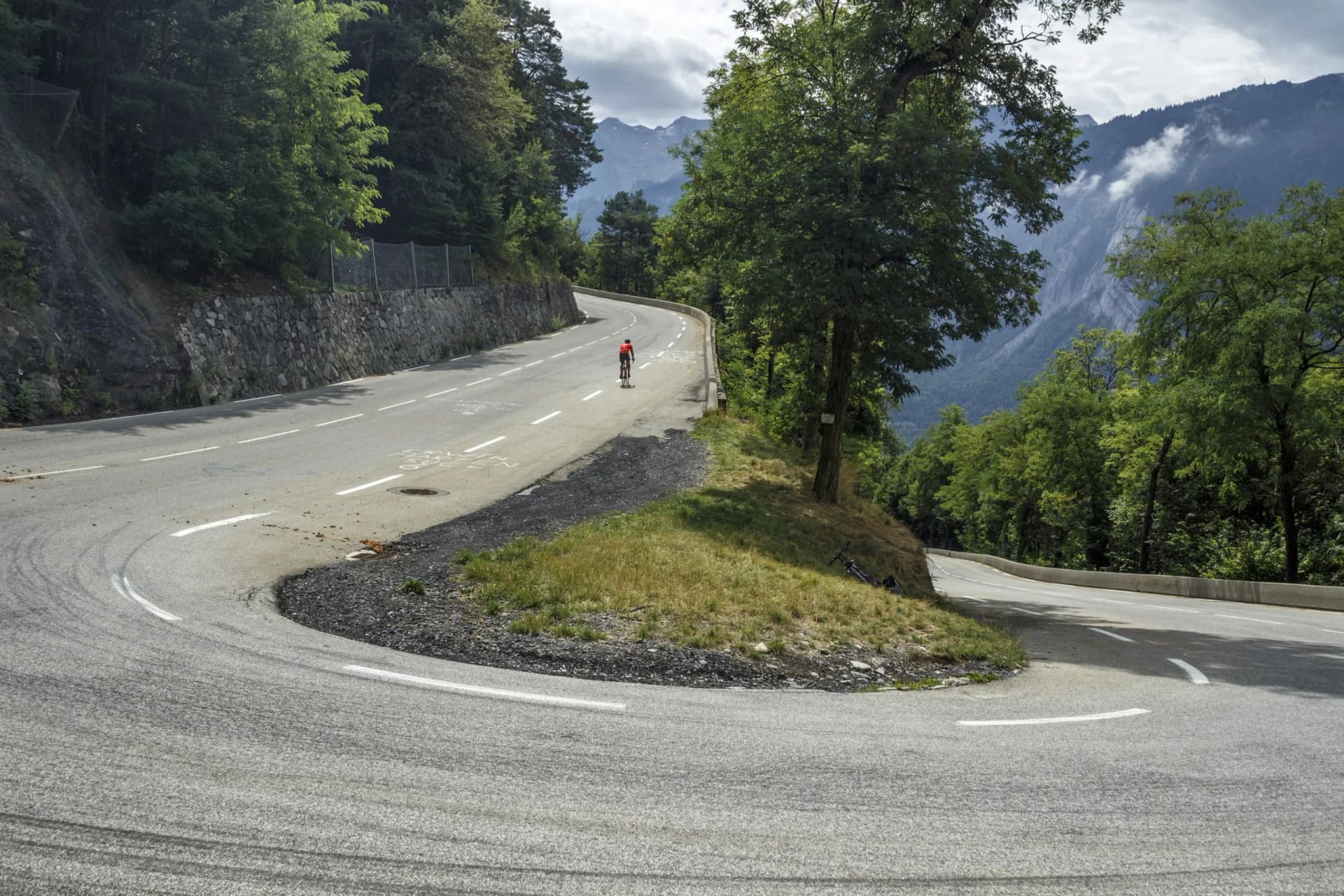 Cyclist ascending winding road with forest and mountains in the background, La Alpe d'Huez, France.