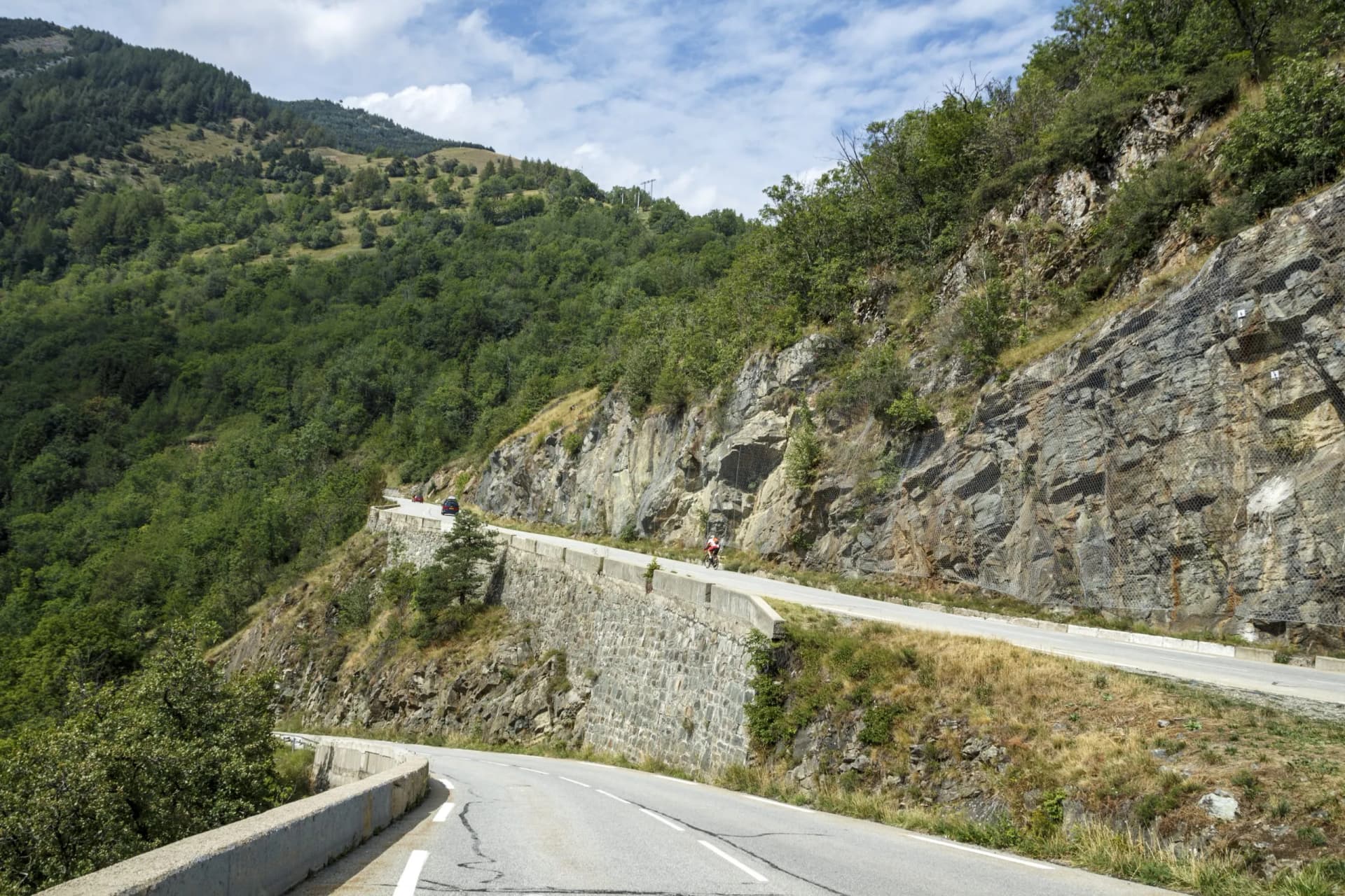 Cyclist ascending steep mountain road with stone retaining wall near La Lpe d'Huez, France.