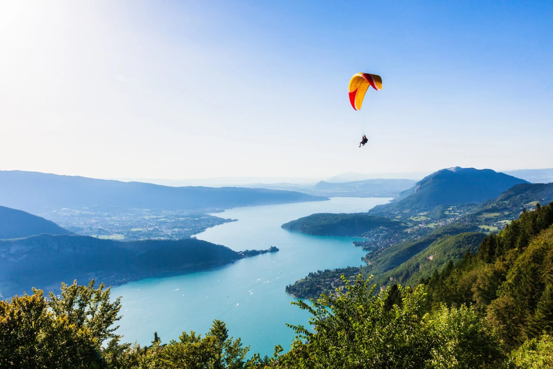 Paraglider soaring over bright blue Annecy Lake and surrounding green mountains.
