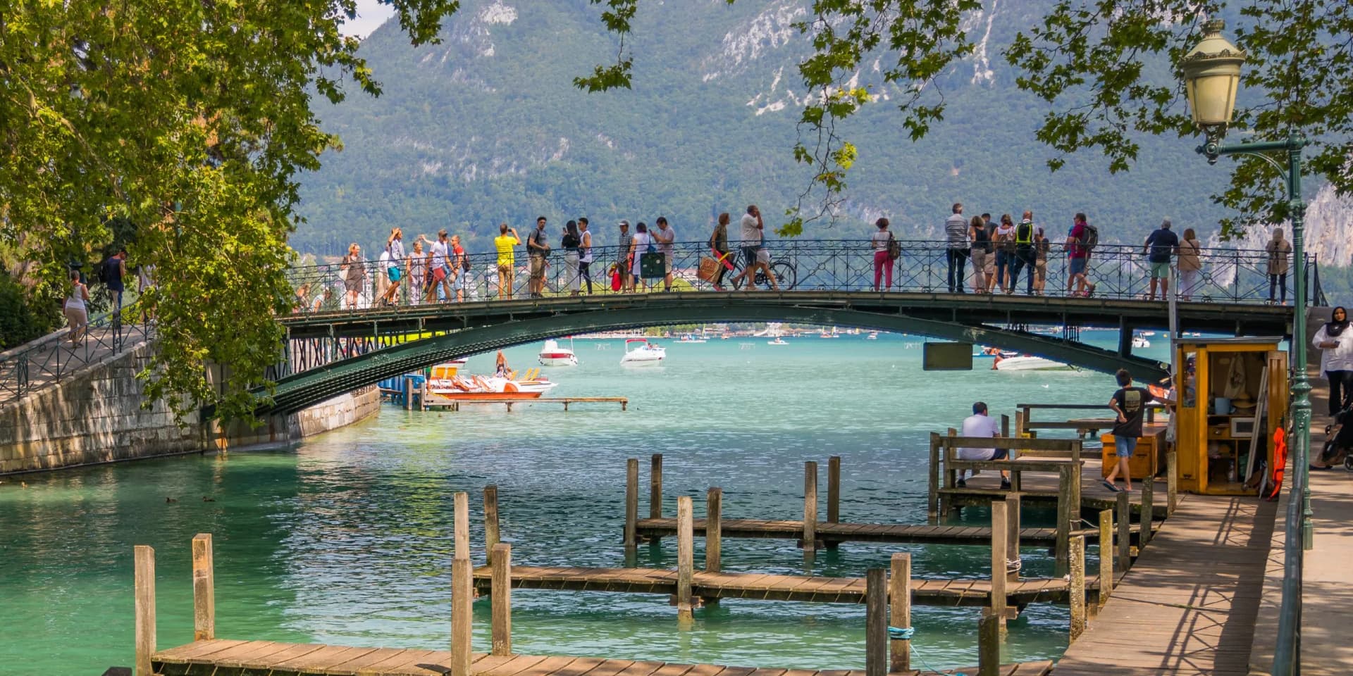 Tourists crossing Pont des Amours bridge over turquoise water with boats and green mountains in Annecy, France.