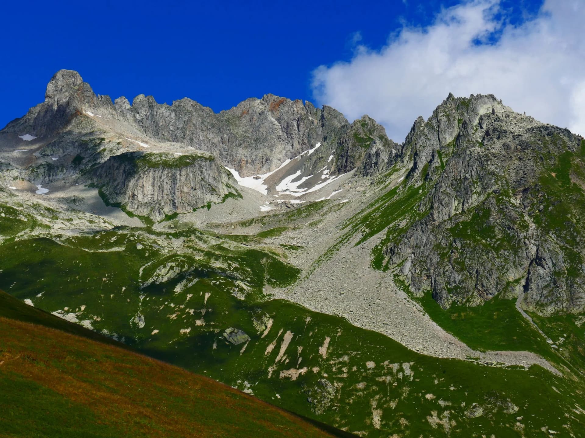 Rugged alpine mountains with green slopes, scree, and patches of snow under a deep blue sky.