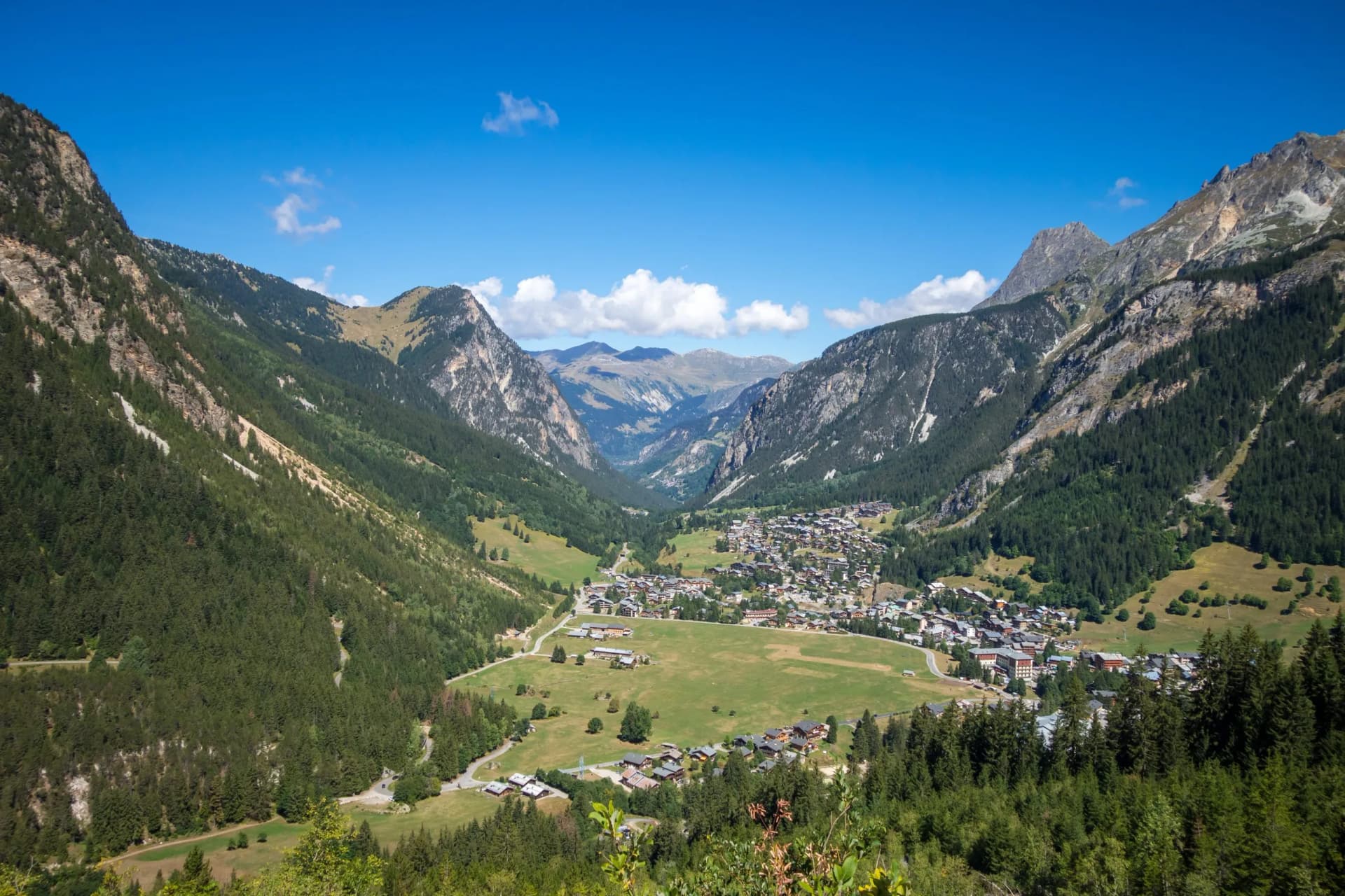 Alpine village nestled in a valley surrounded by steep, forested mountains under a bright blue sky.