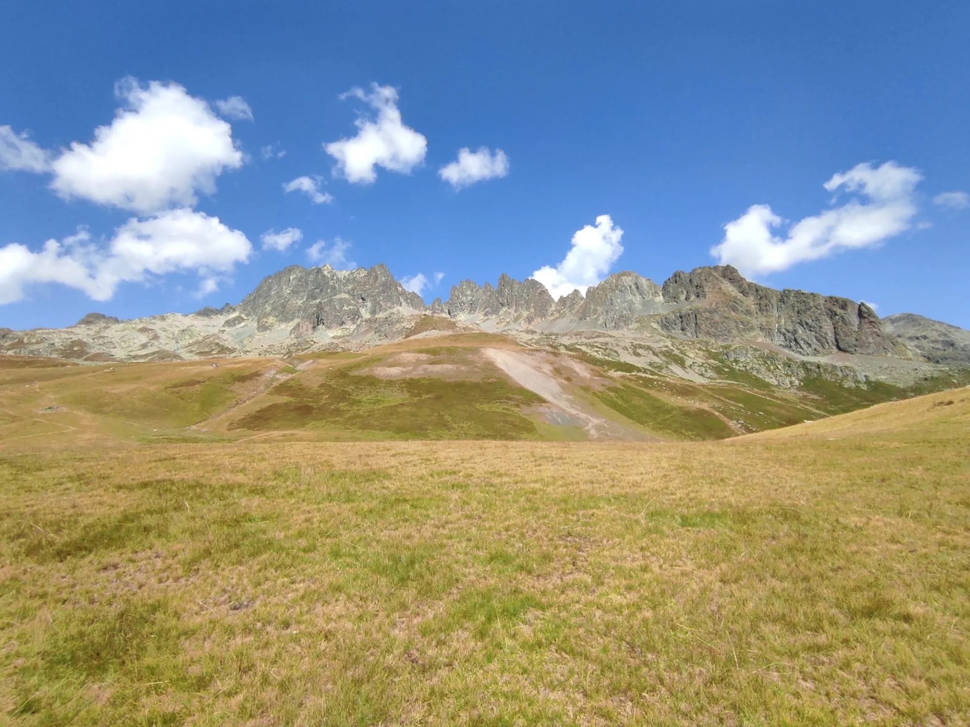 Grassy summer alpine meadow leading to rugged mountain peaks under a blue sky with white clouds near Col du Glandon.