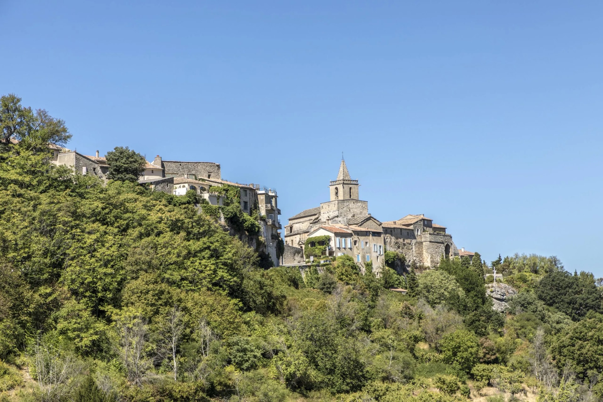 Stone village perched on a densely wooded hillside under a clear blue sky in Venasque.