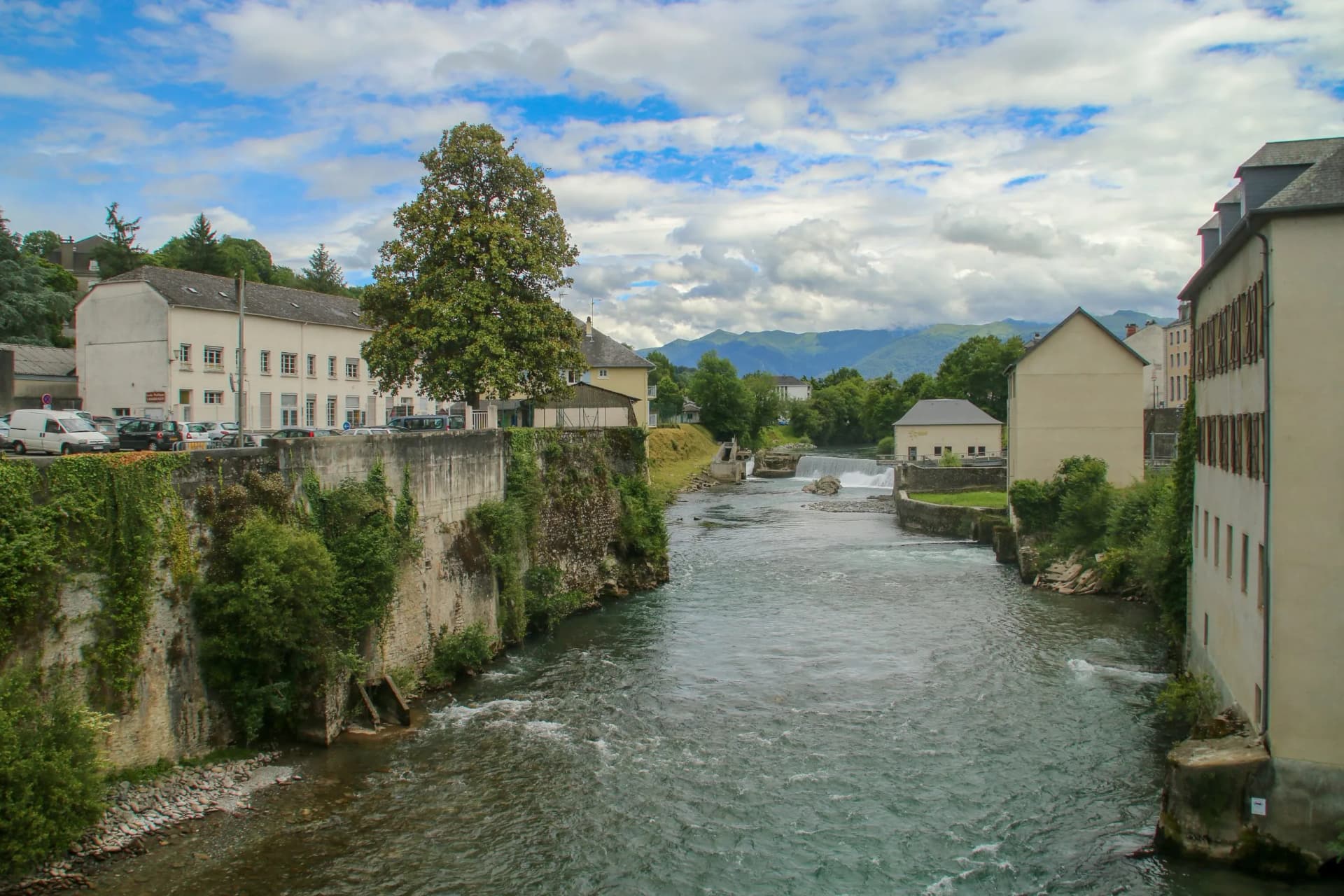 River flowing through Oloron-Sainte-Marie with stone banks, buildings, and mountains under a cloudy sky.