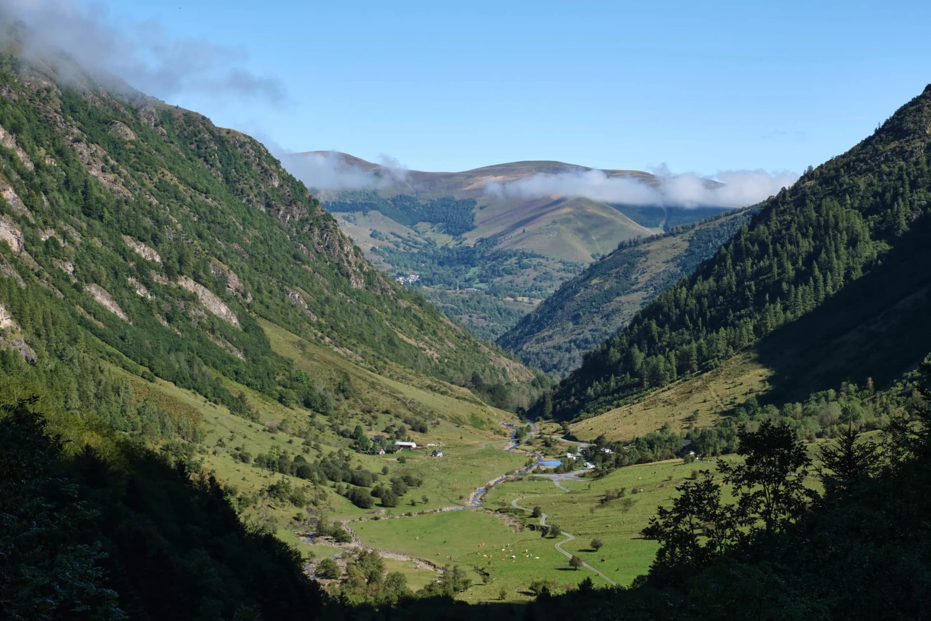 Mountain valley with green slopes, winding river, small buildings, and low clouds in Les Granges d'Astau, France.