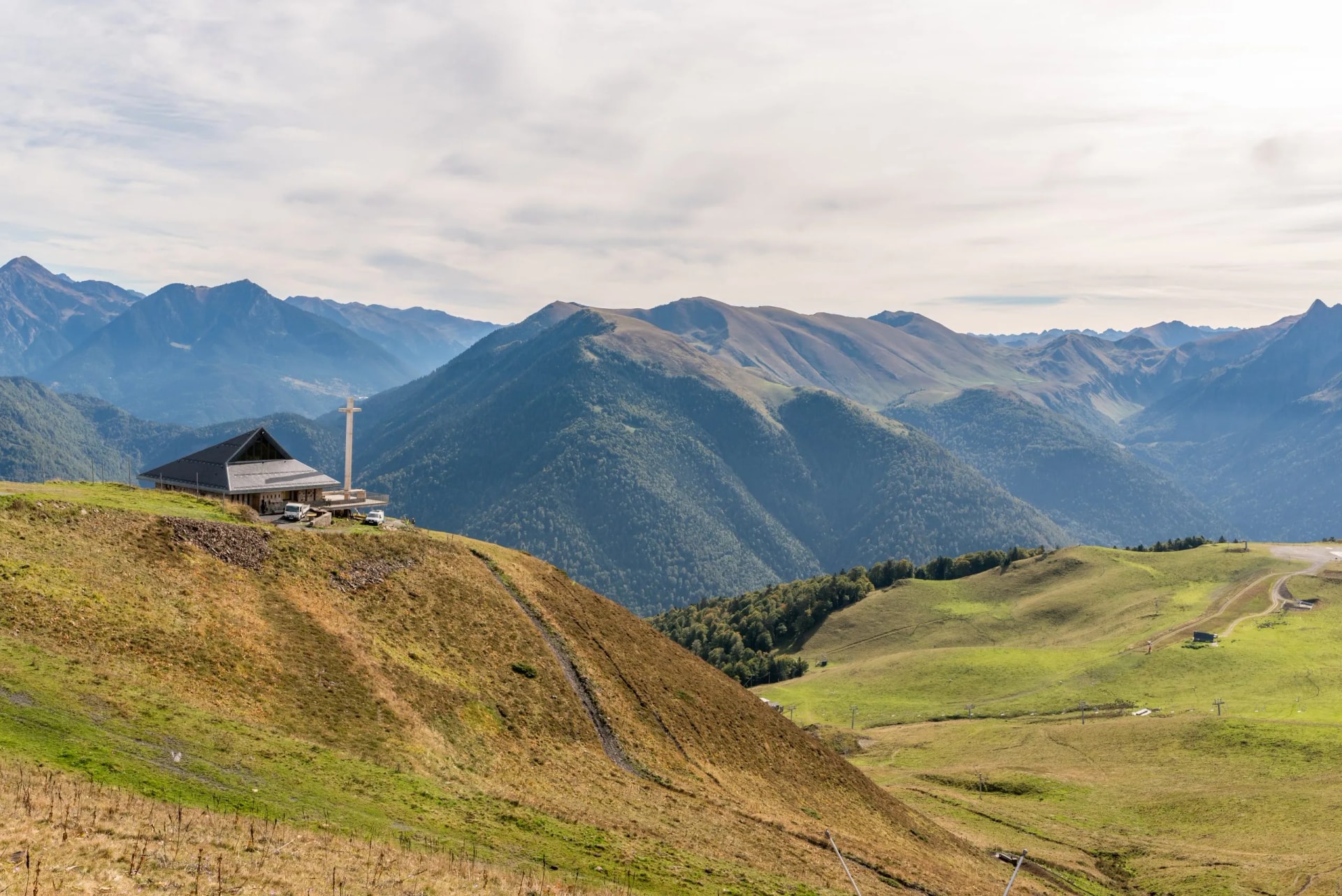 Mountain view from Luchon resort with a modern building and large cross overlooking forested Pyrenees peaks.