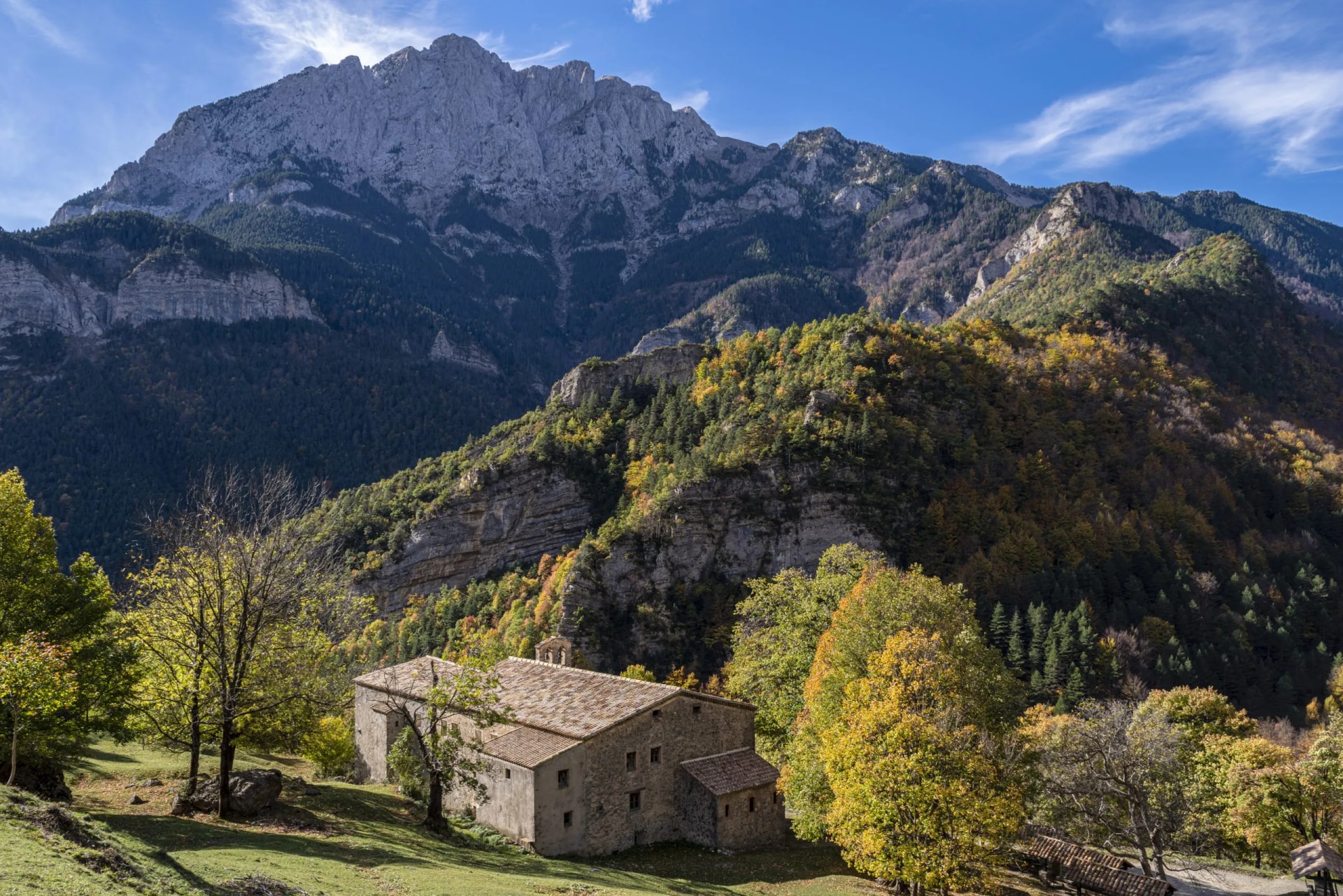 Stone sanctuary below massive rocky mountain in El Pedraforca, Catalonia, with autumn trees.