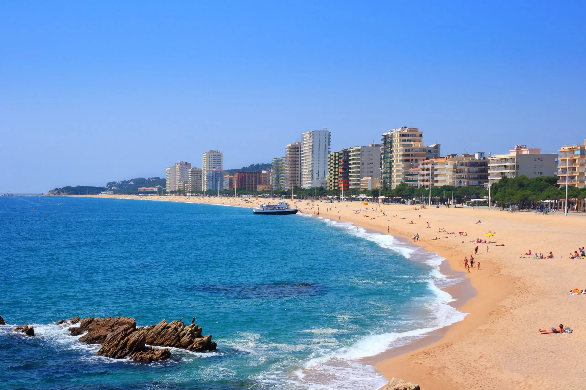 Beach with sunbathers, boat, and high-rise buildings in Platja d'Aro, Costa Brava, Spain.