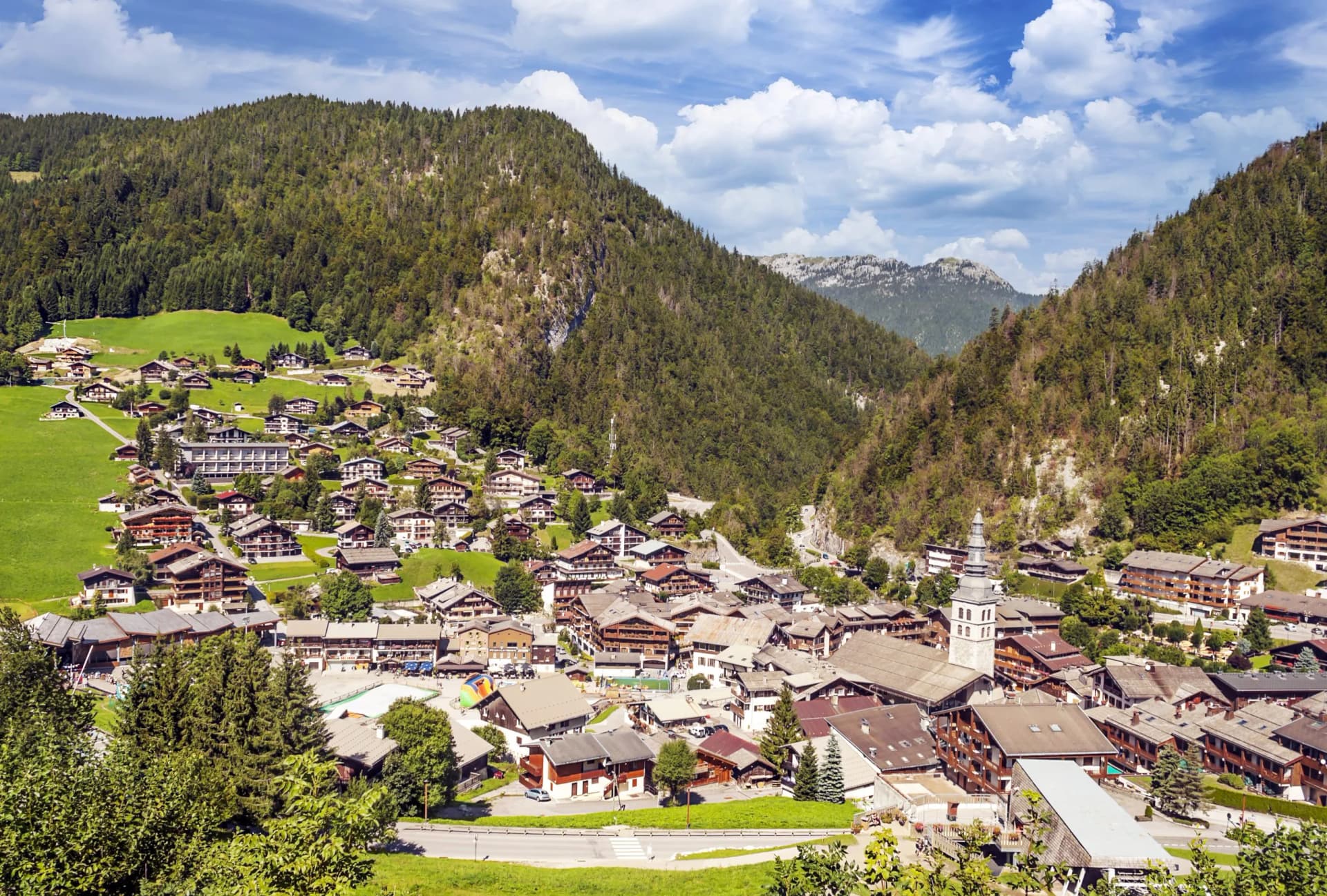 Alpine village nestled in a valley with wooden chalets and a church tower under a cloudy sky.