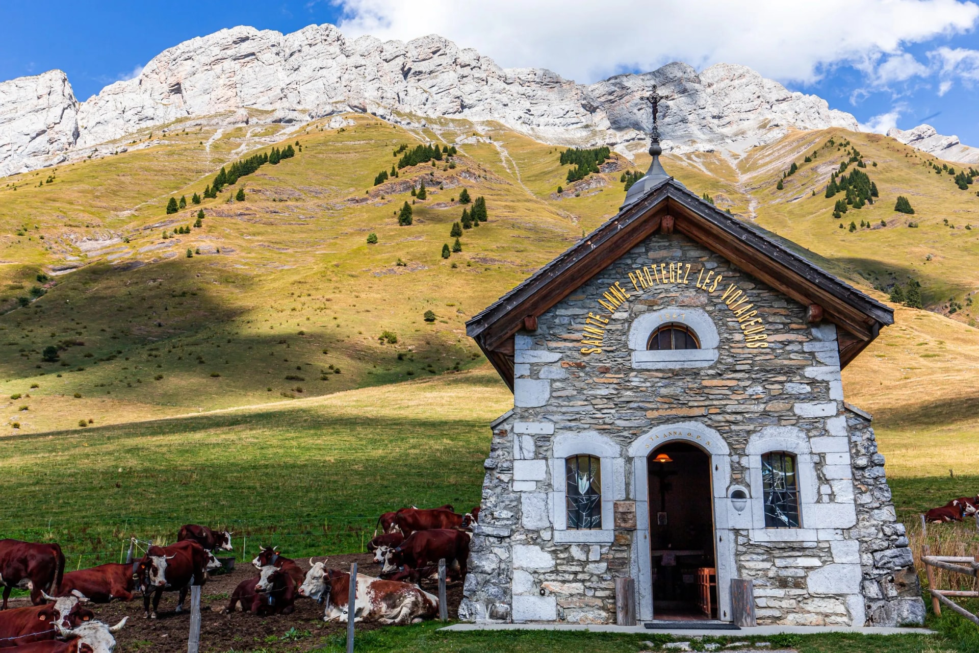 Cows grazing near a stone chapel below rocky mountains near La Clusaz, Haute-Savoie, France.