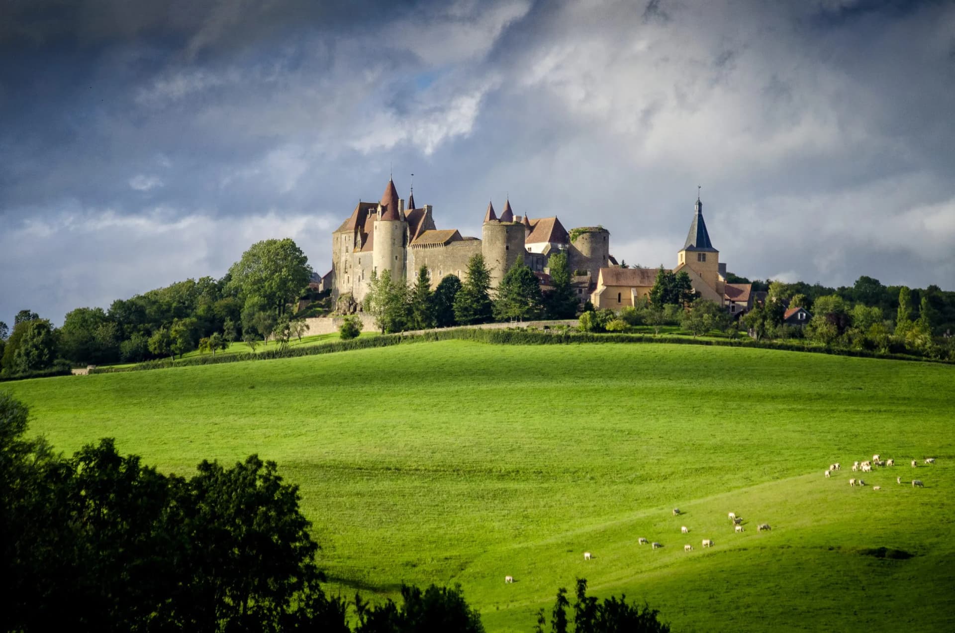 Châteauneuf fortress above bright green hillside with grazing sheep under dramatic sky.