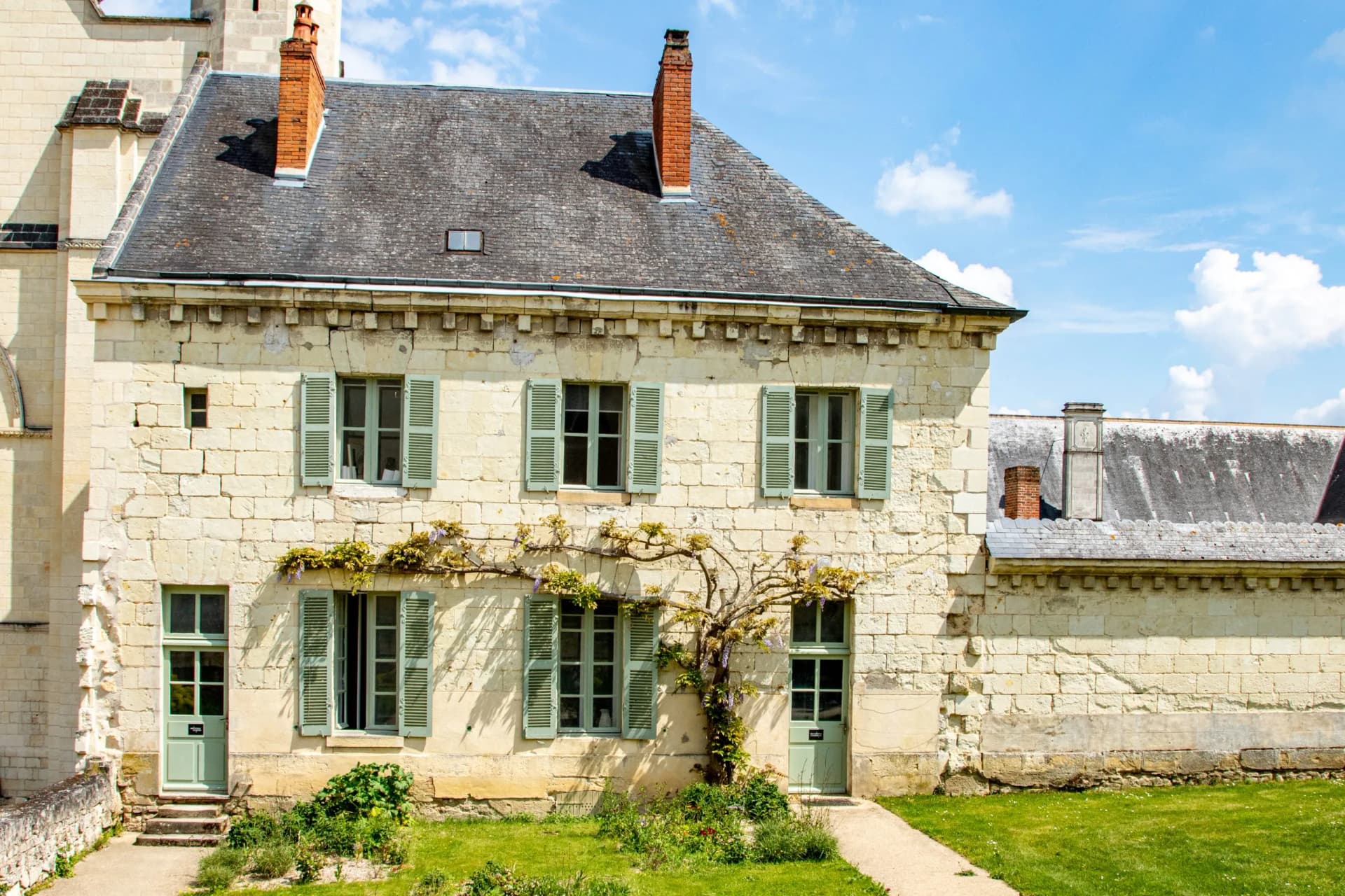 Stone building with light green shutters and climbing wisteria under a blue sky at Fontevraud Abbey.