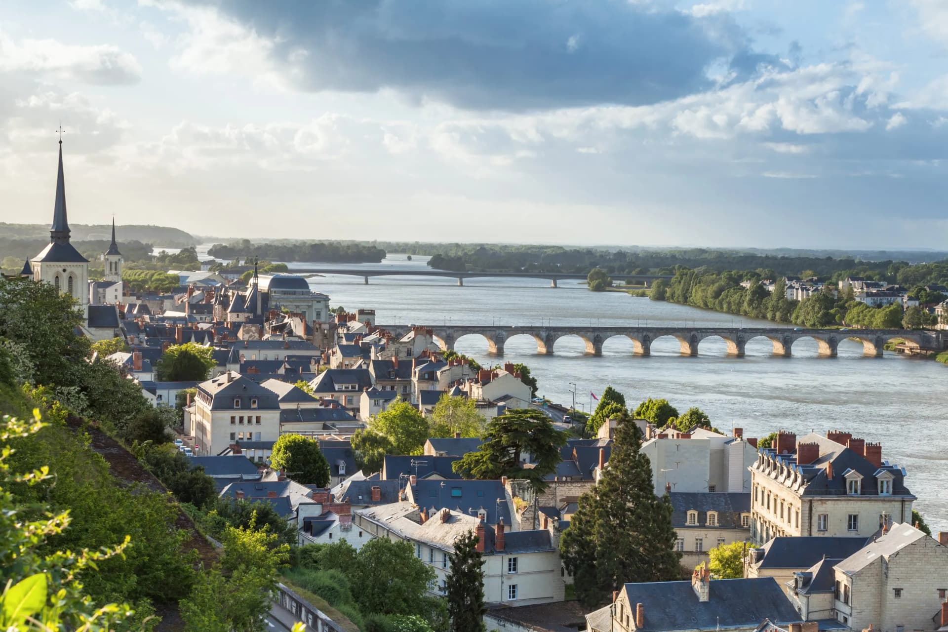 View of Saumur, France, with historic town, Loire River, and arched bridge under cloudy sky.